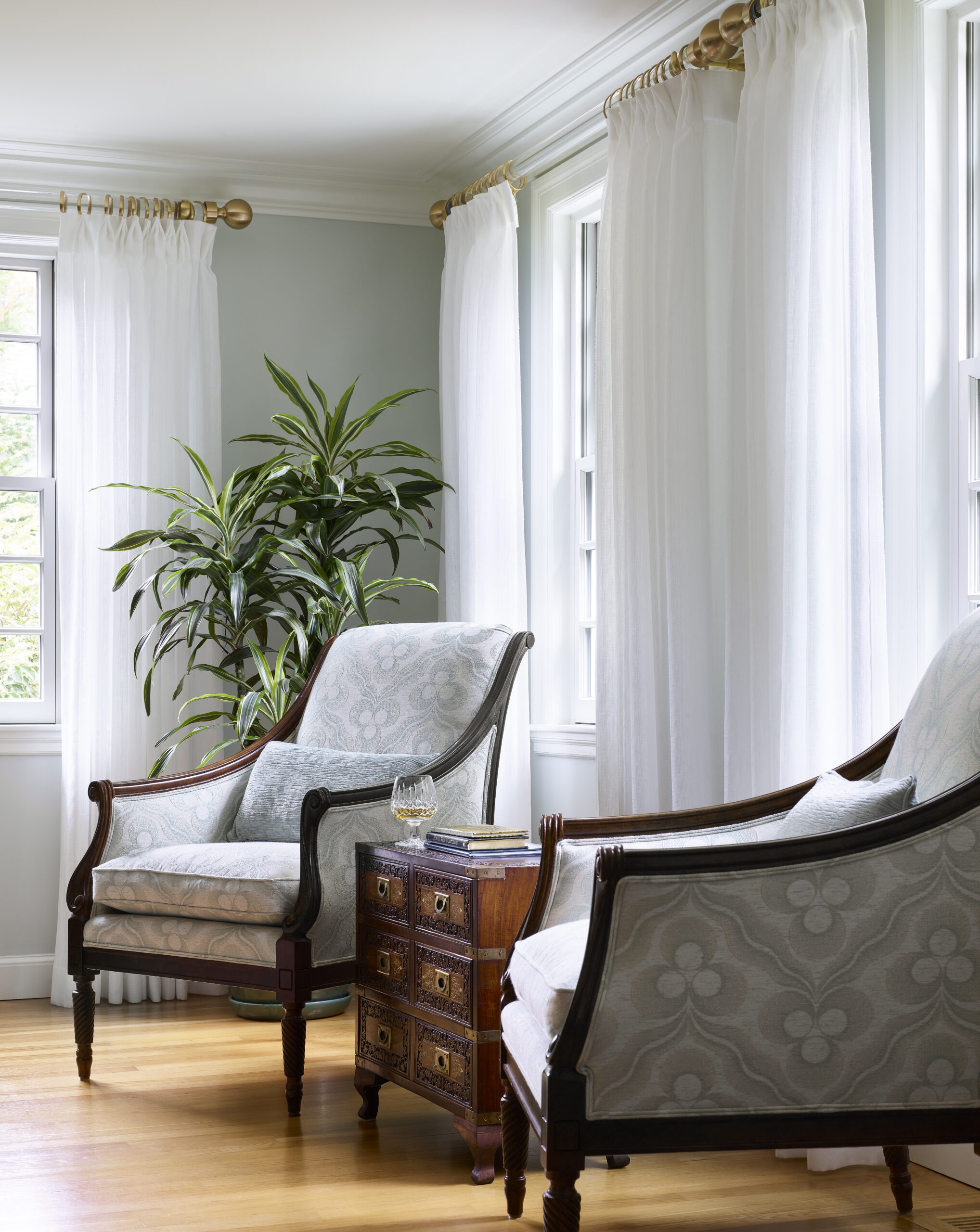 Two upholstered armchairs and a small wooden side table with glass sit by large windows with sheer white curtains, reflecting luxury interior design. A tall green plant stands in the corner, adding elegance to the serene setting.