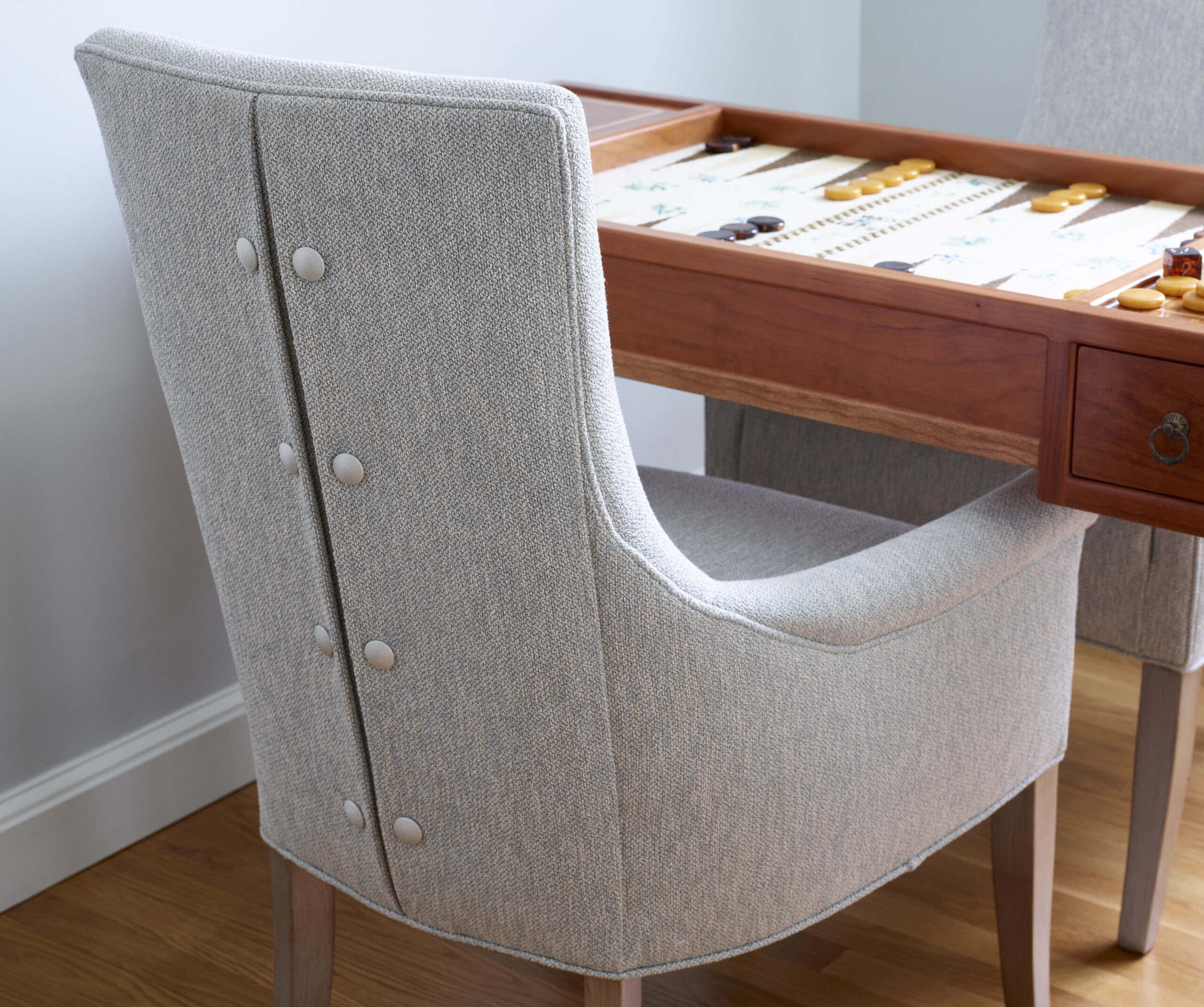 A light gray upholstered chair with button details is positioned at a wooden table with a backgammon board set up for play, reflecting the elegance of luxury interior design.