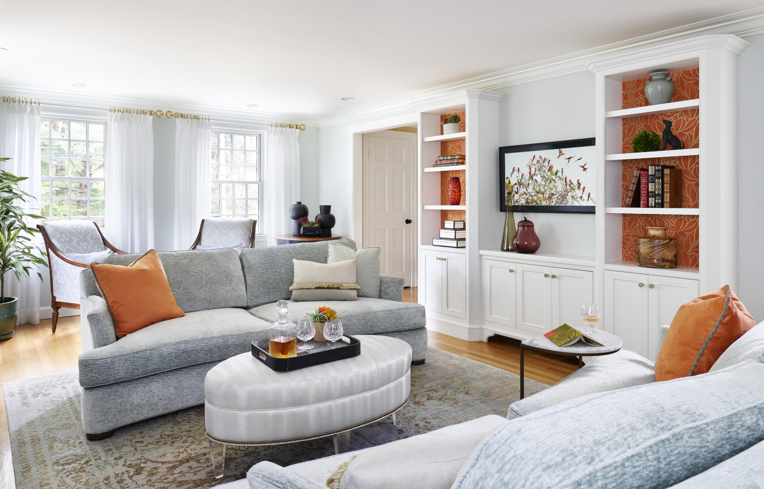 Bright living room with light blue sectional sofa, orange throw pillows, white ottoman, built-in shelves, large windows with sheer curtains, and decorative accents—styled by an Interior Designer Massachusetts for a touch of luxury interior design.