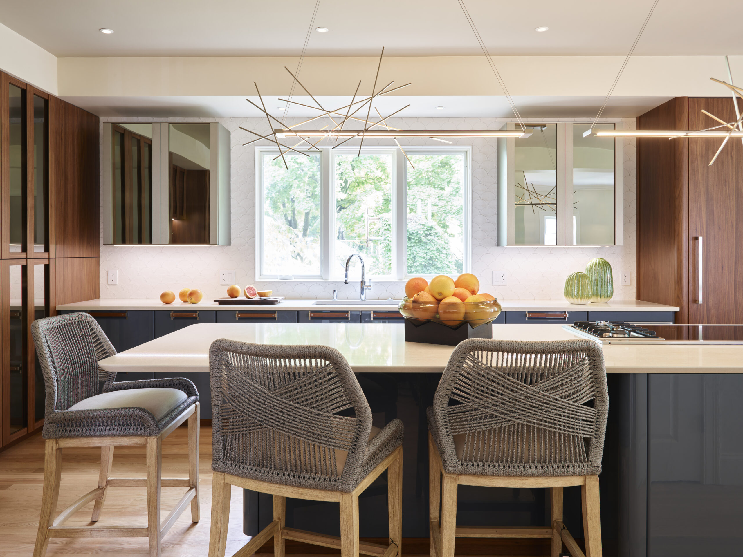 Modern kitchen with wood cabinets, a large island with woven bar stools, pendant lights, and a bowl of oranges on the counter in front of a window—showcasing luxury interior design by an expert Interior Designer Massachusetts.
