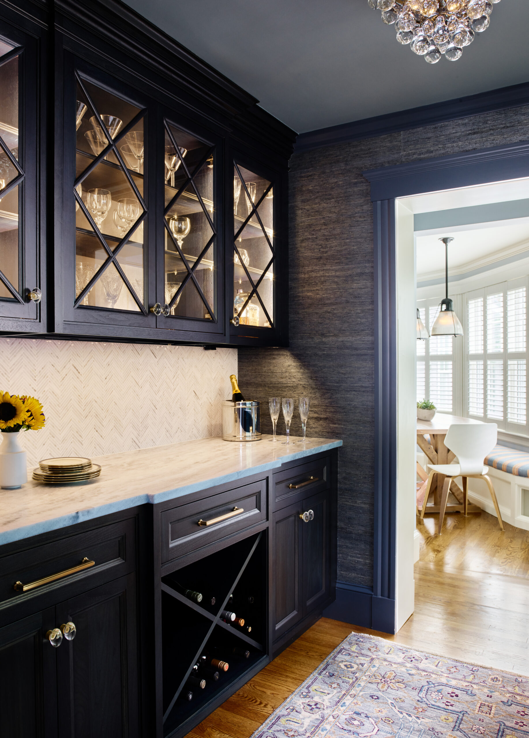 A butler's pantry with dark cabinets, glass-front uppers showcasing glassware, a marble countertop, and a wine rack reflects luxury interior design as it leads to a bright dining area—perfect inspiration from an interior designer Massachusetts.