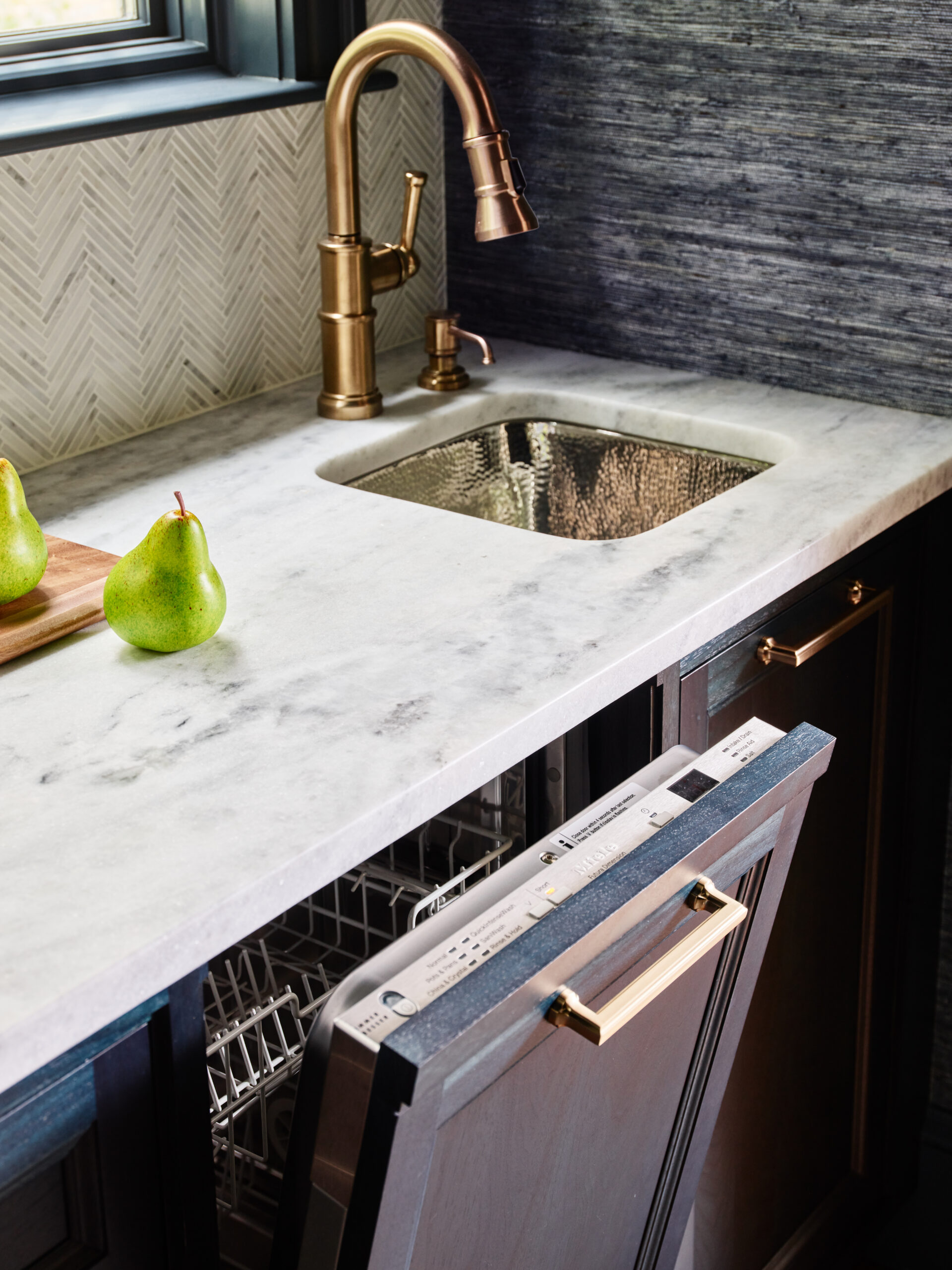 A small sink with a brass faucet is set in a marble countertop, reflecting luxury interior design. Two pears rest on a cutting board nearby, and a dishwasher below is open with the rack partially visible.
