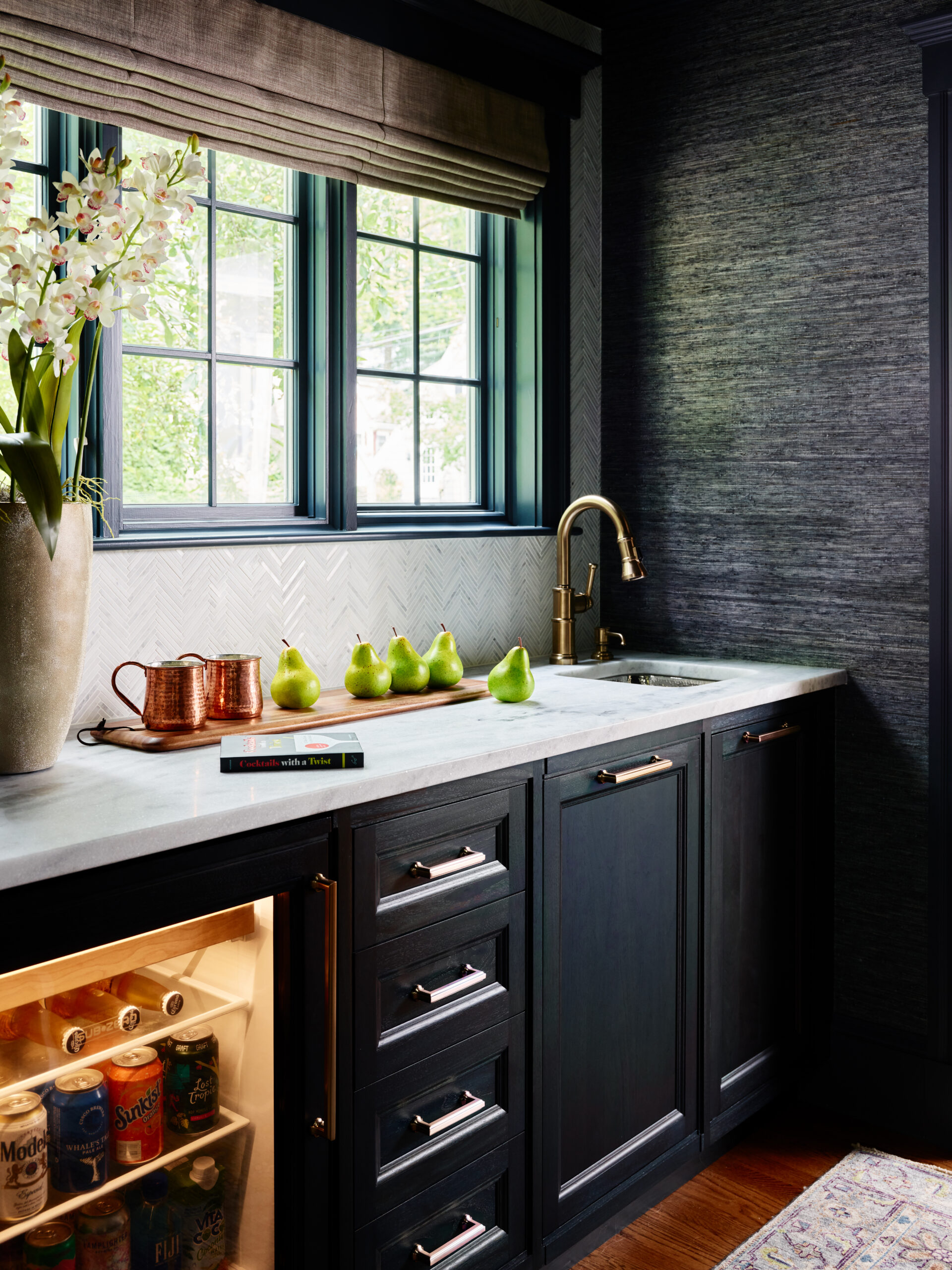 A kitchen counter with a sink, brass faucet, vase of flowers, copper mug, and pears exudes Luxury Interior Design. Below, a mini fridge stocked with drinks is set in dark cabinetry for a refined touch.