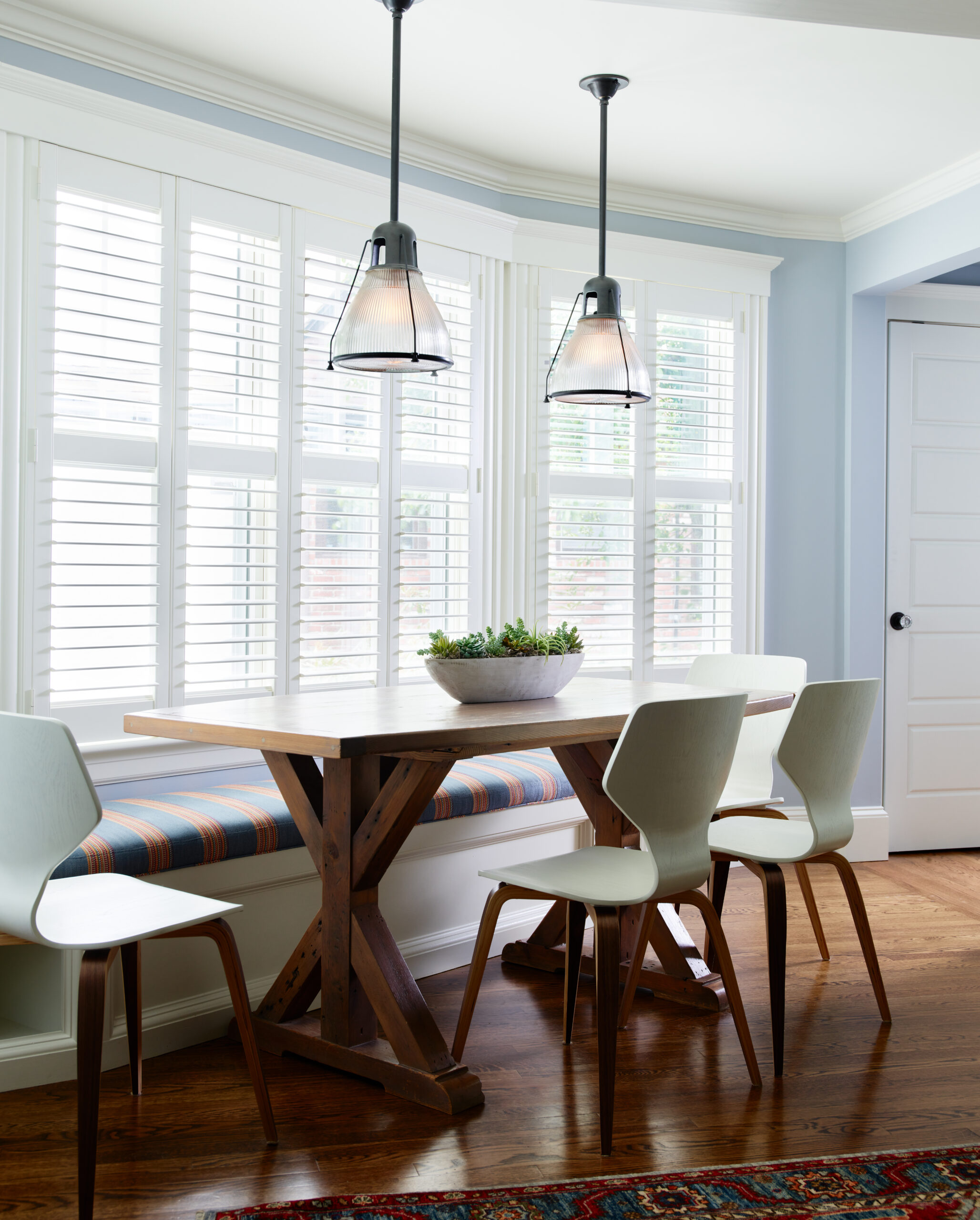 A dining nook by an Interior Designer Massachusetts features a wooden table, four white chairs, a built-in bench with striped cushion, two pendant lights, and large windows with white shutters for a touch of luxury interior design.