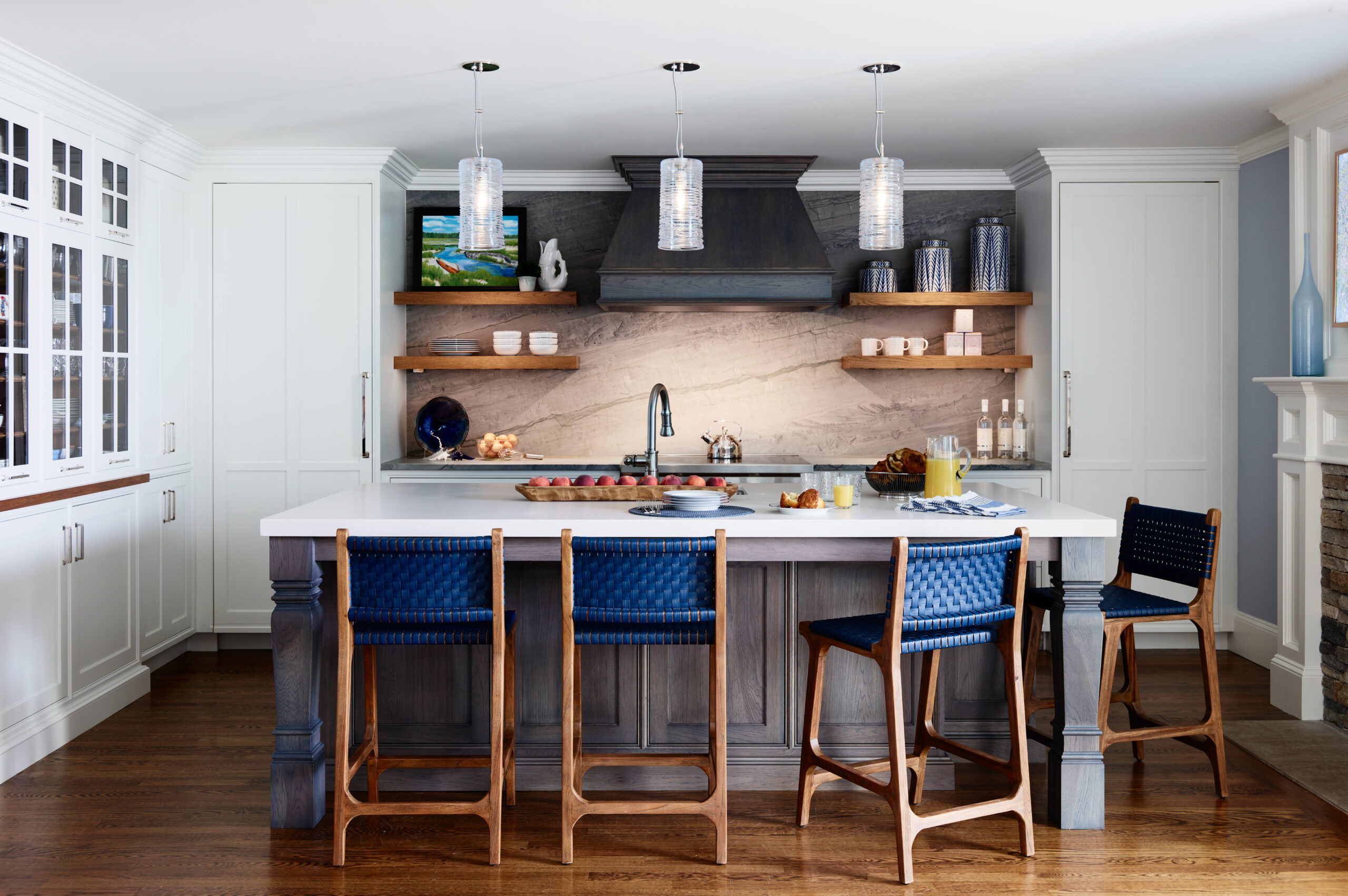 Modern kitchen by a top Interior Designer in Massachusetts, featuring white cabinets, a marble backsplash, blue woven chairs at the island, pendant lights, and floating shelves—an inviting space reflecting luxury interior design.