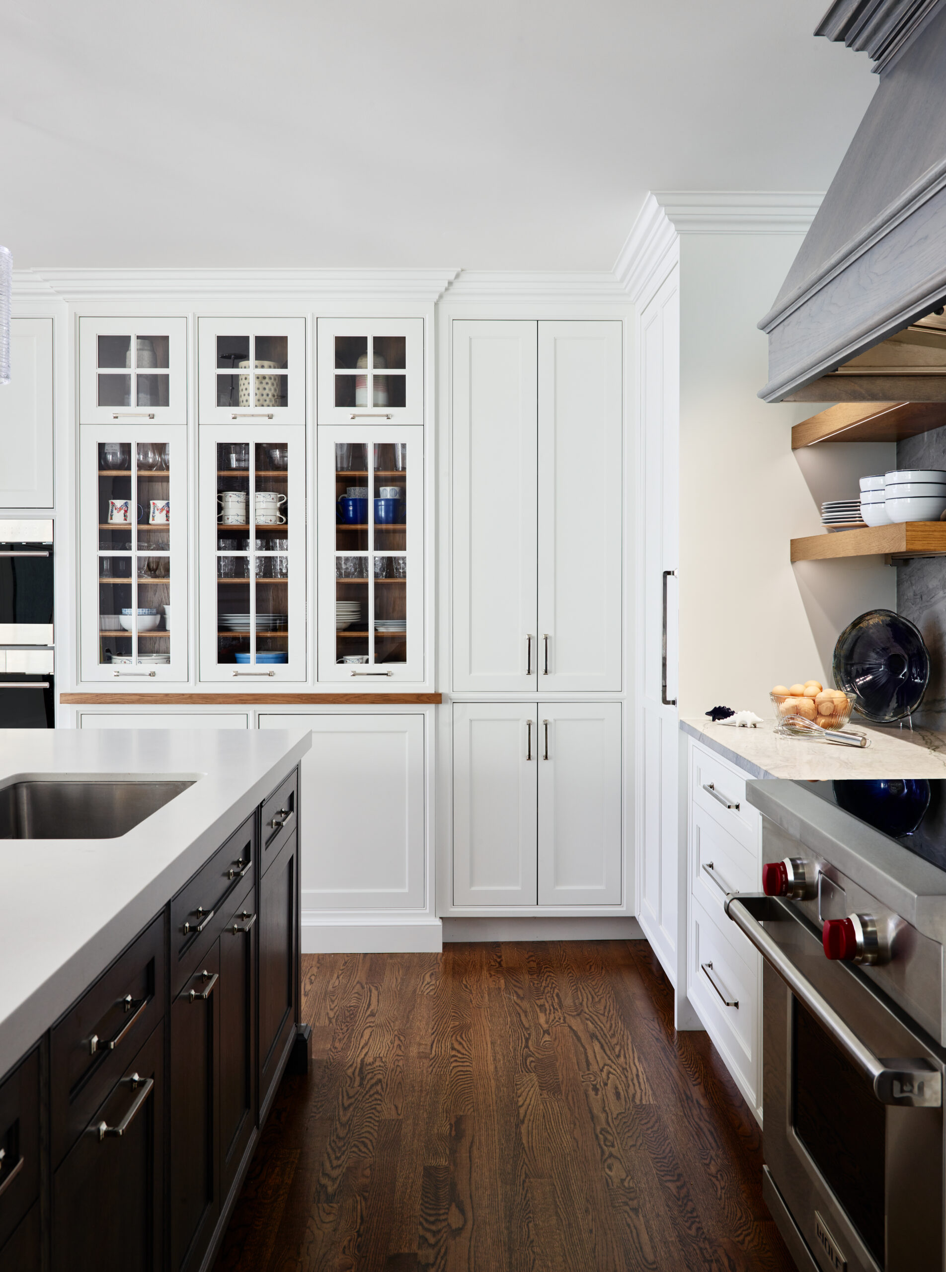 Modern kitchen with white cabinets, glass-front uppers displaying dishes, a dark wood island with a sink, and stainless steel appliances on dark hardwood floors—an example of luxury interior design by an Interior Designer Massachusetts.