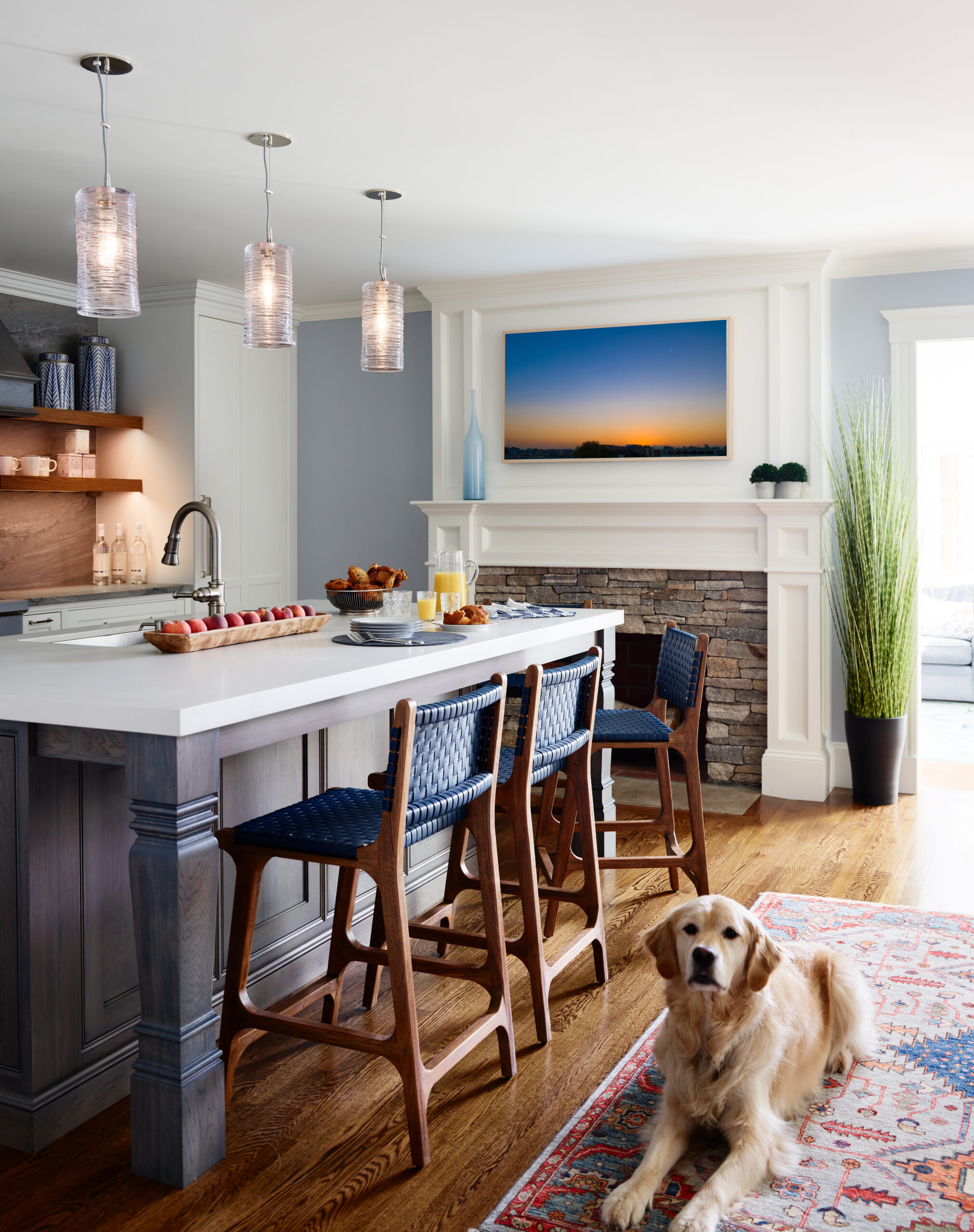 Modern kitchen with a large island, three blue stools, pendant lights, food on the counter, a fireplace with art above, a tall plant, and a golden retriever lying on a patterned rug—luxury interior design by an Interior Designer in Massachusetts.