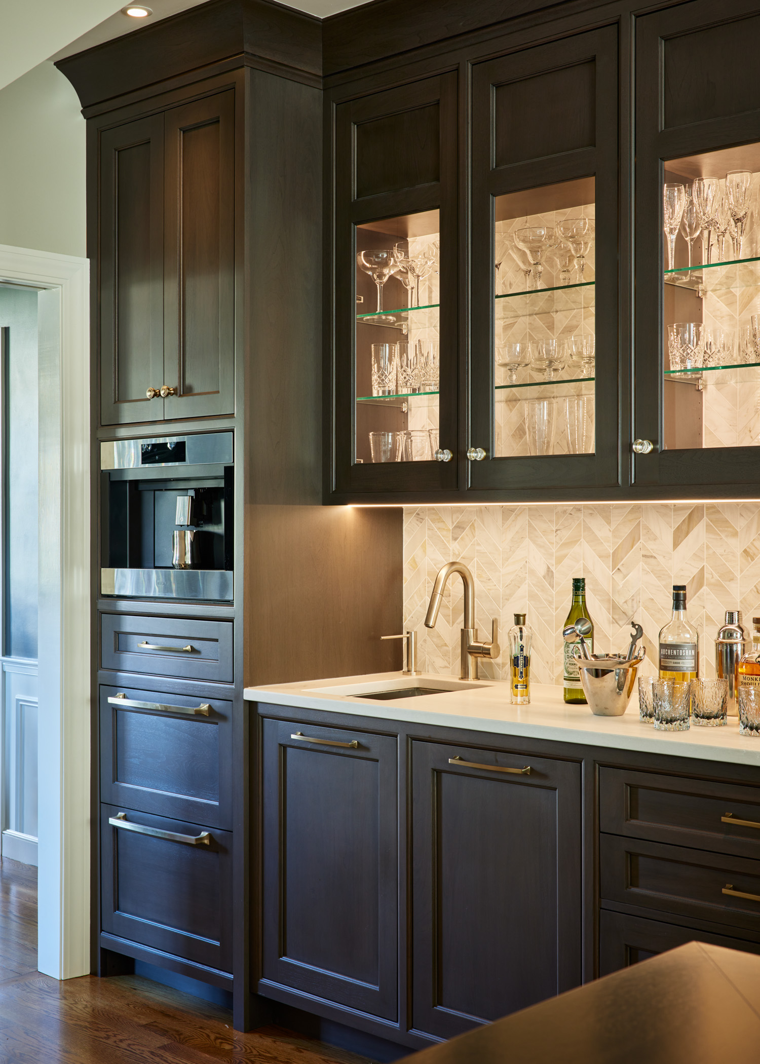 Modern kitchen bar area with dark wood cabinets, glass-front upper shelves displaying glassware, built-in oven, sink, and neatly arranged bar tools—showcasing luxury interior design inspired by top Interior Designer Massachusetts.