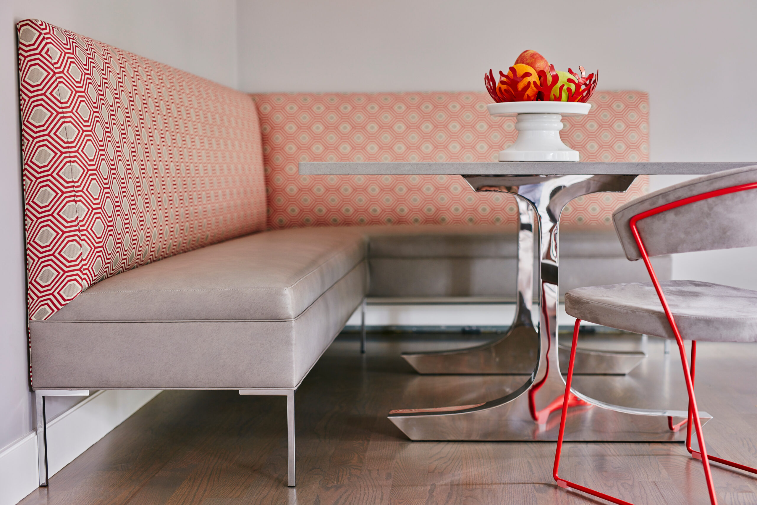 Modern dining nook by an Interior Designer Massachusetts, featuring an upholstered corner bench, geometric patterns, metal table, gray chairs with red legs, and a white fruit bowl centerpiece on a wood floor—Luxury Interior Design at its finest.