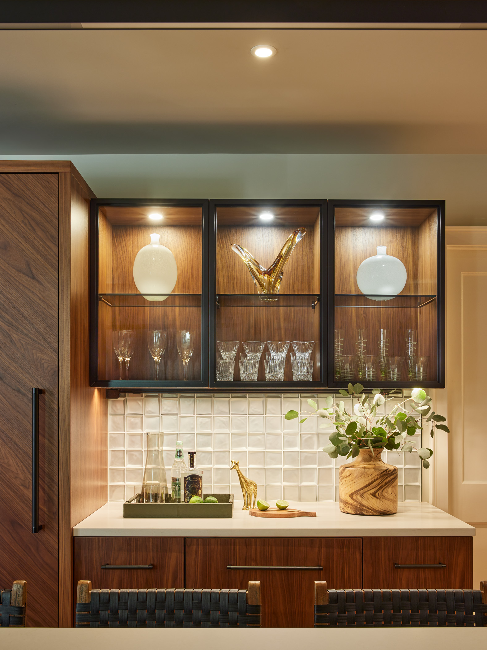 A modern kitchen bar area with wood cabinets, glassware, decorative vases, a gold animal figurine, and a plant in a wooden vase on a white countertop showcases luxury interior design at its finest.