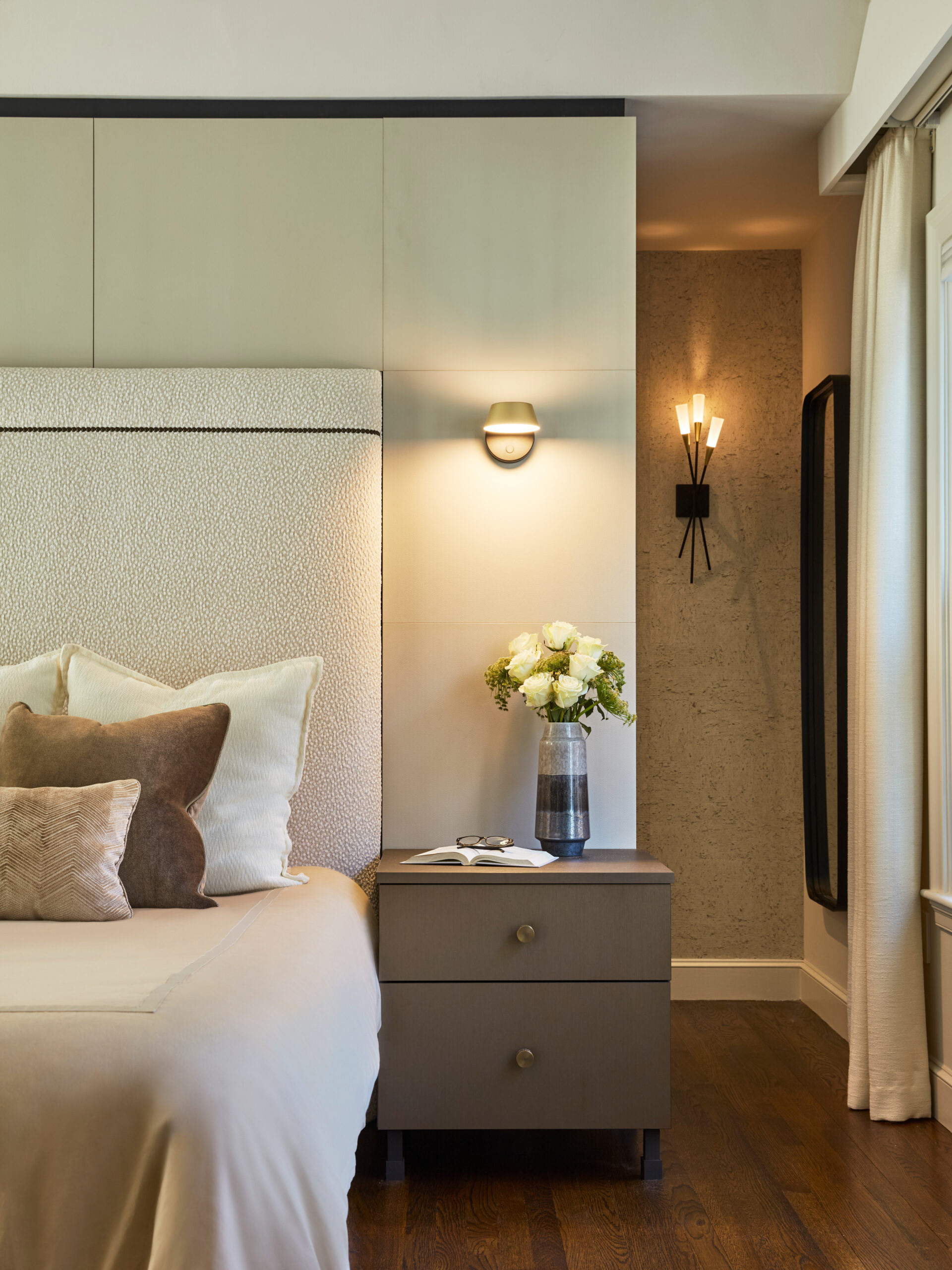 Modern bedroom with a bed, neutral bedding, decorative pillows, nightstand with a vase of white flowers, wall sconces, and a window with light curtains—an elegant example of luxury interior design by an interior designer in Massachusetts.