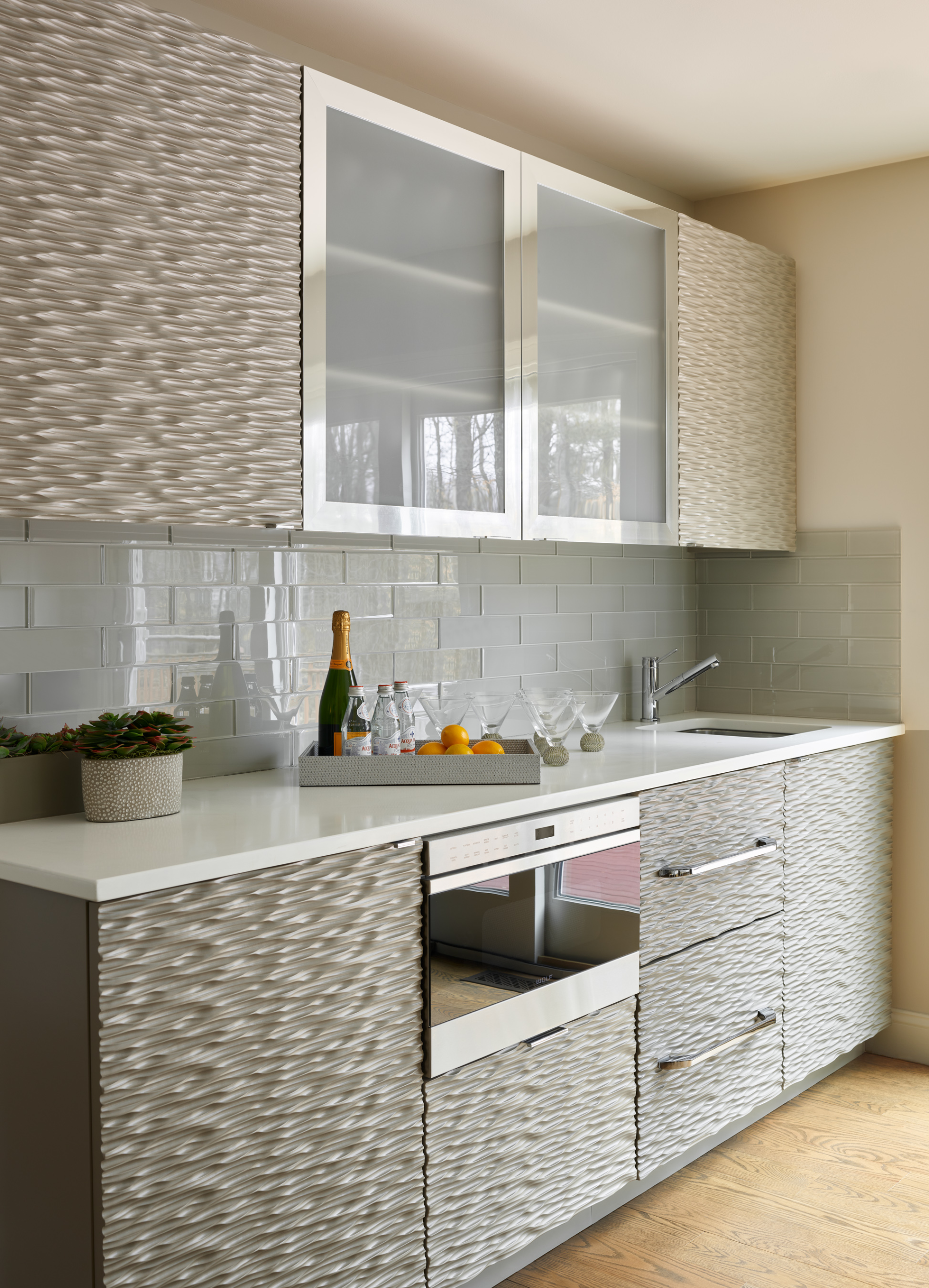 Modern kitchen by an Interior Designer Massachusetts, featuring textured cabinets, glass-front uppers, glossy subway tile backsplash, and a countertop set with champagne, glasses, lemons, and a potted plant—perfect for luxury interior design lovers.