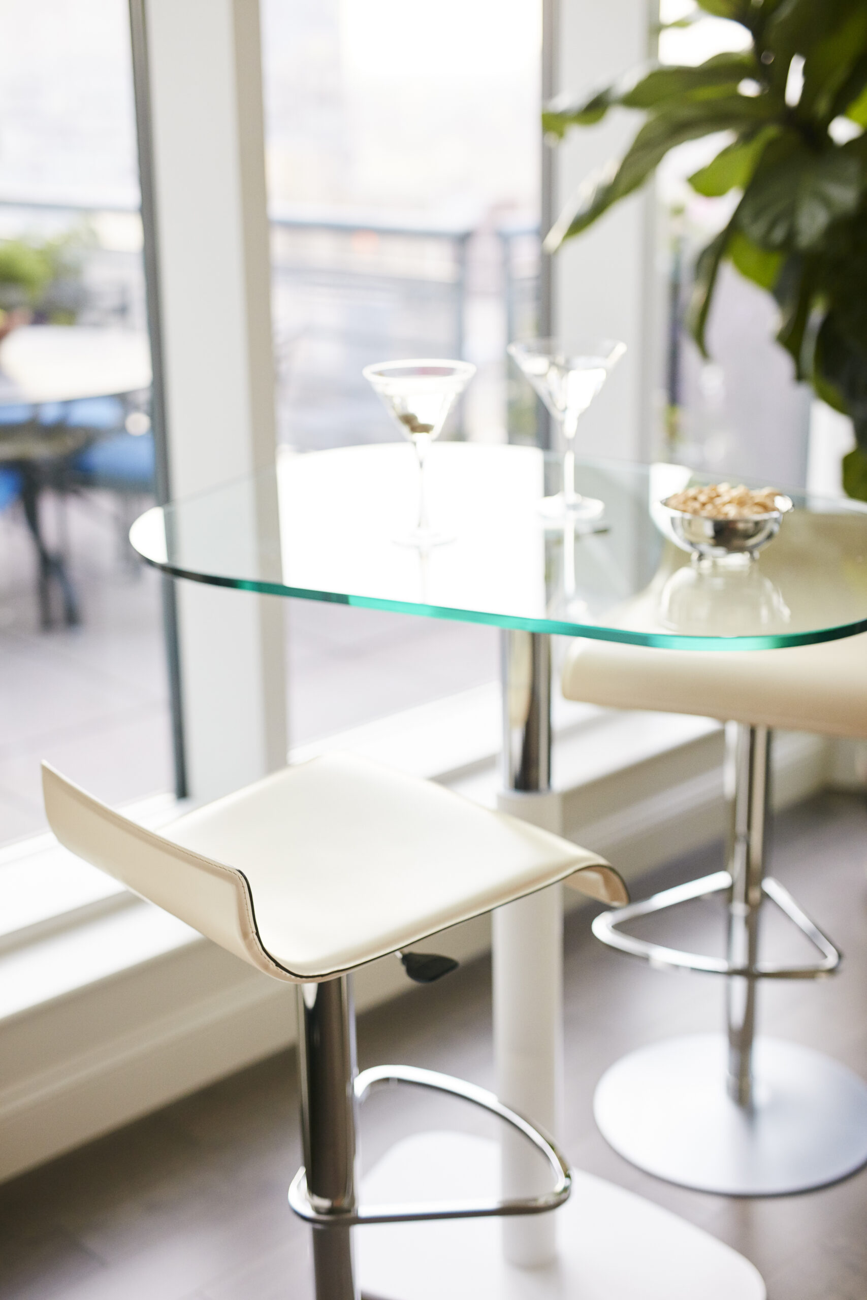 A glass-top table with two martini glasses and a bowl of snacks, next to two white bar stools by large windows and a potted plant—perfect for showcasing luxury interior design flair.