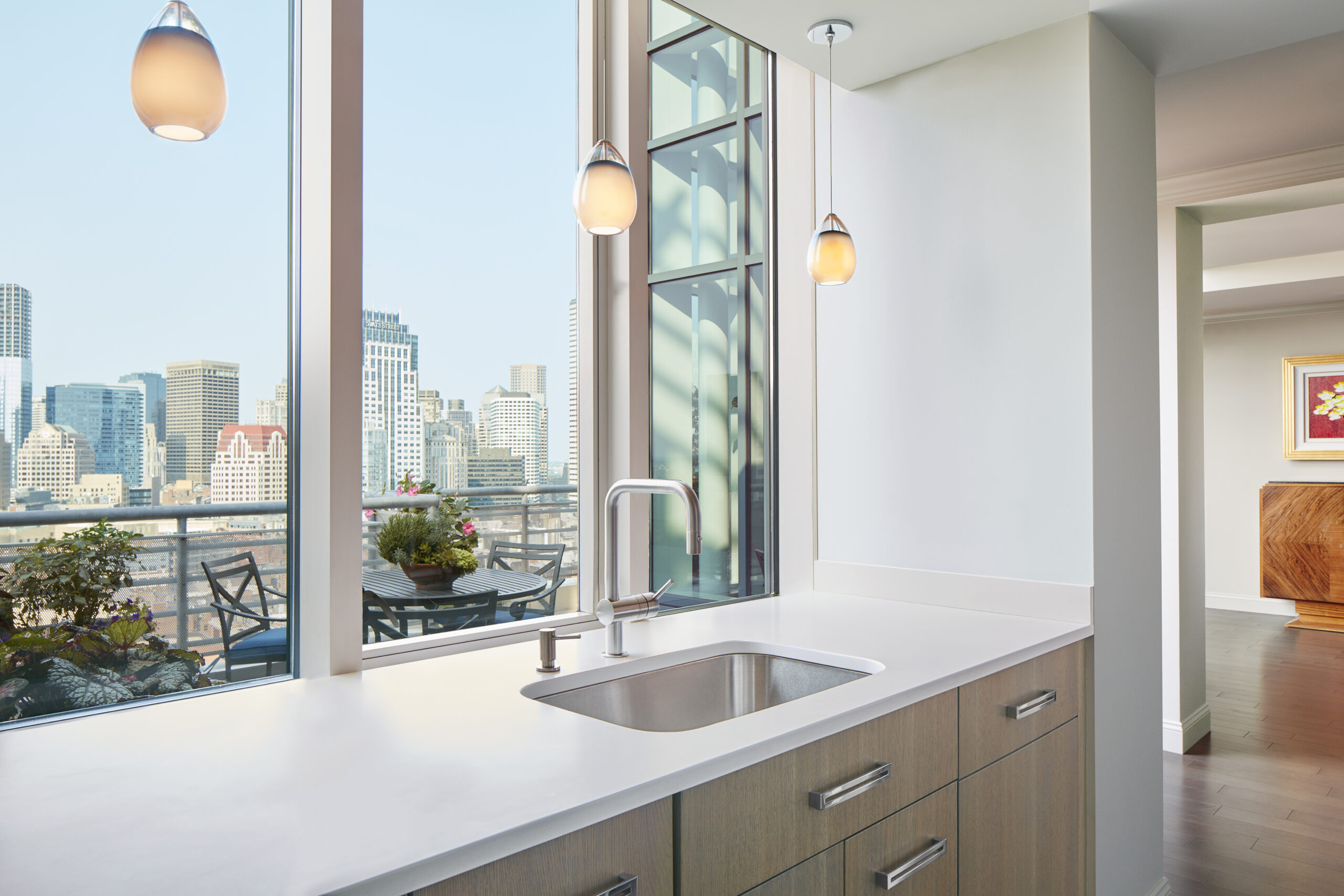 Modern kitchen sink with a sleek faucet, light wood cabinets, and hanging pendant lights, showcasing luxury interior design by an expert interior designer in Massachusetts, overlooking a cityscape through large windows.