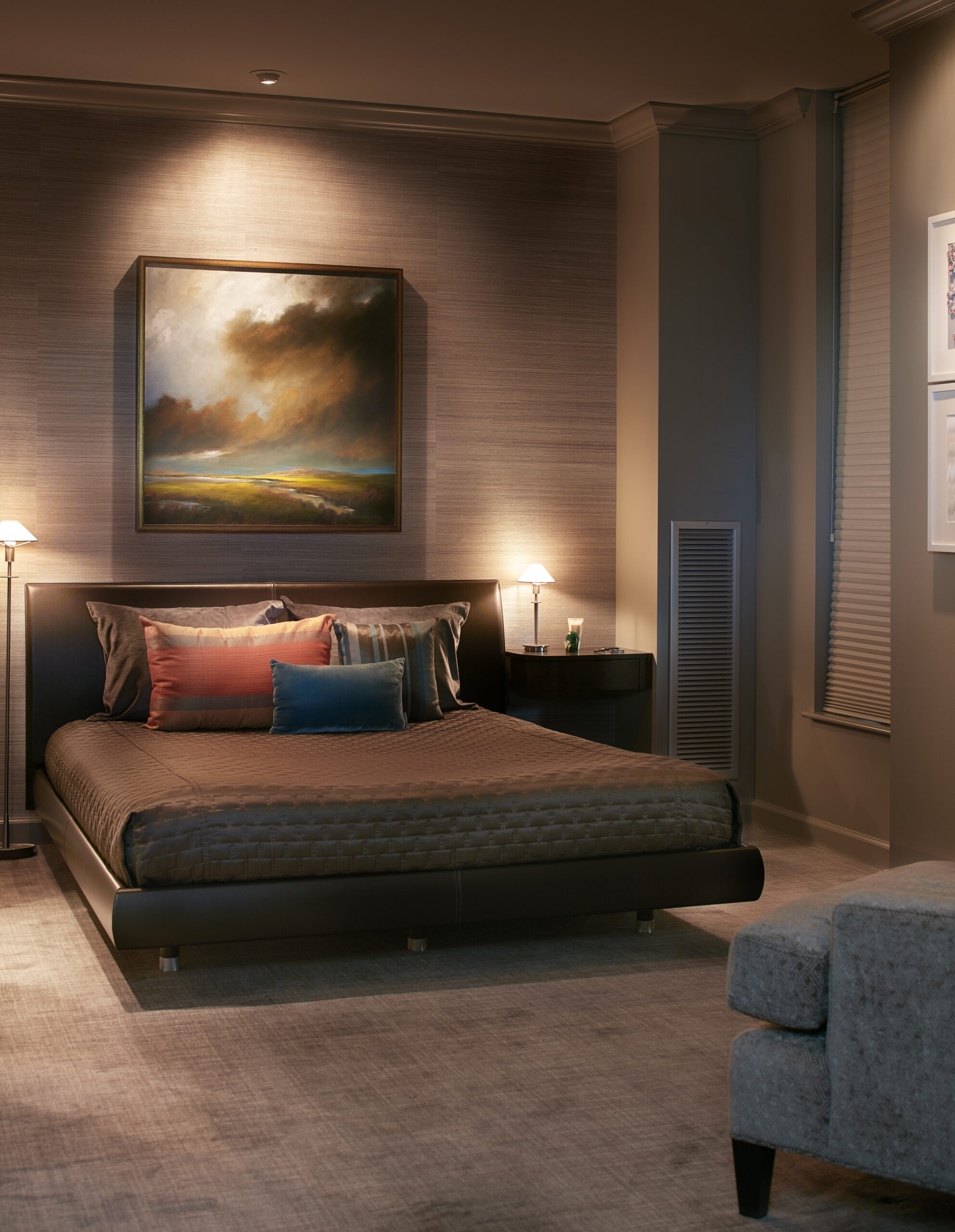 A modern bedroom with a low dark bed, assorted pillows, two bedside lamps, a large wall painting, and neutral décor—perfectly capturing the elegance of luxury interior design by an expert interior designer in Massachusetts.