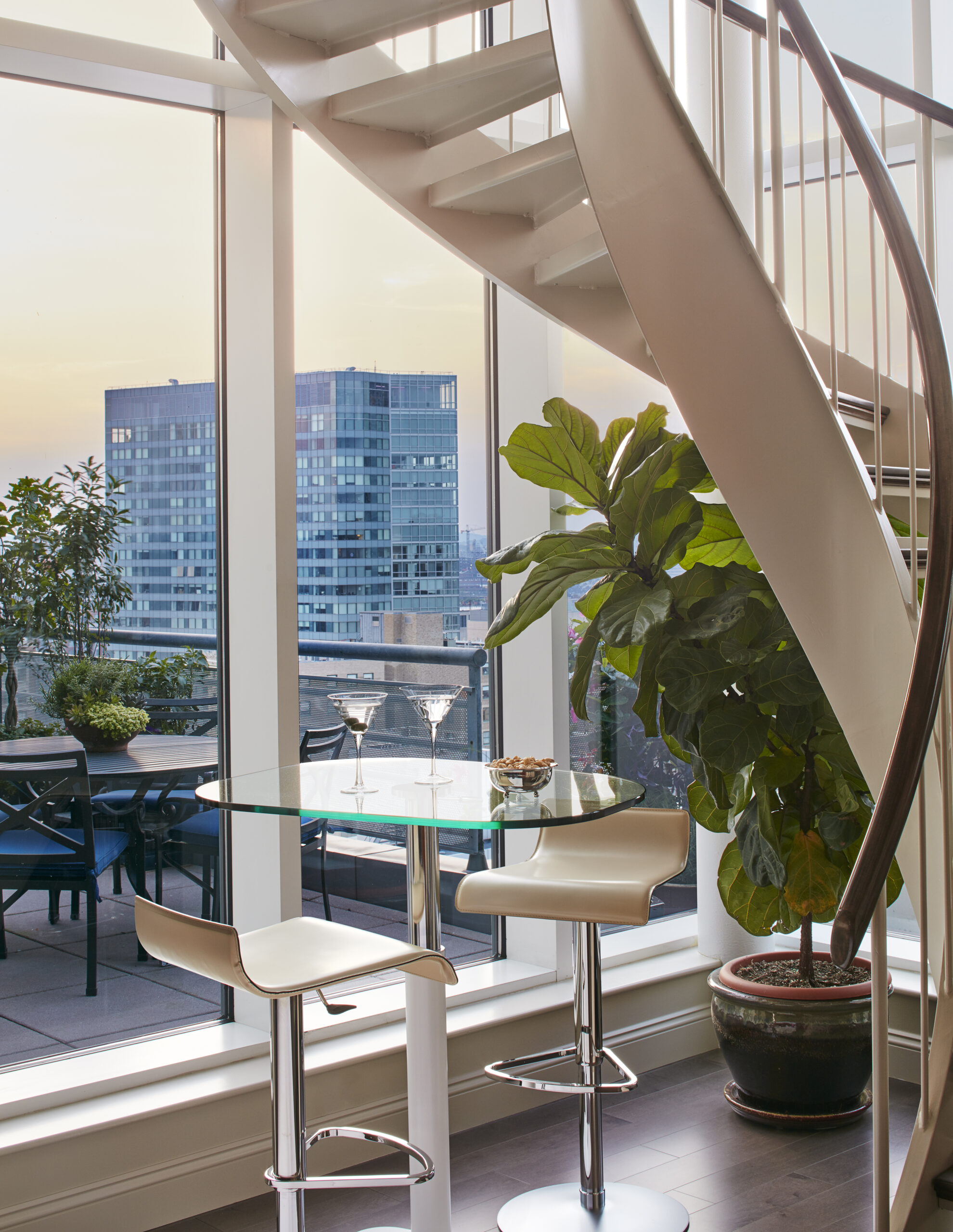 A modern glass table with two bar stools sits by a spiral staircase in a bright room with large windows, showcasing luxury interior design and city views, crafted by an expert interior designer in Massachusetts.