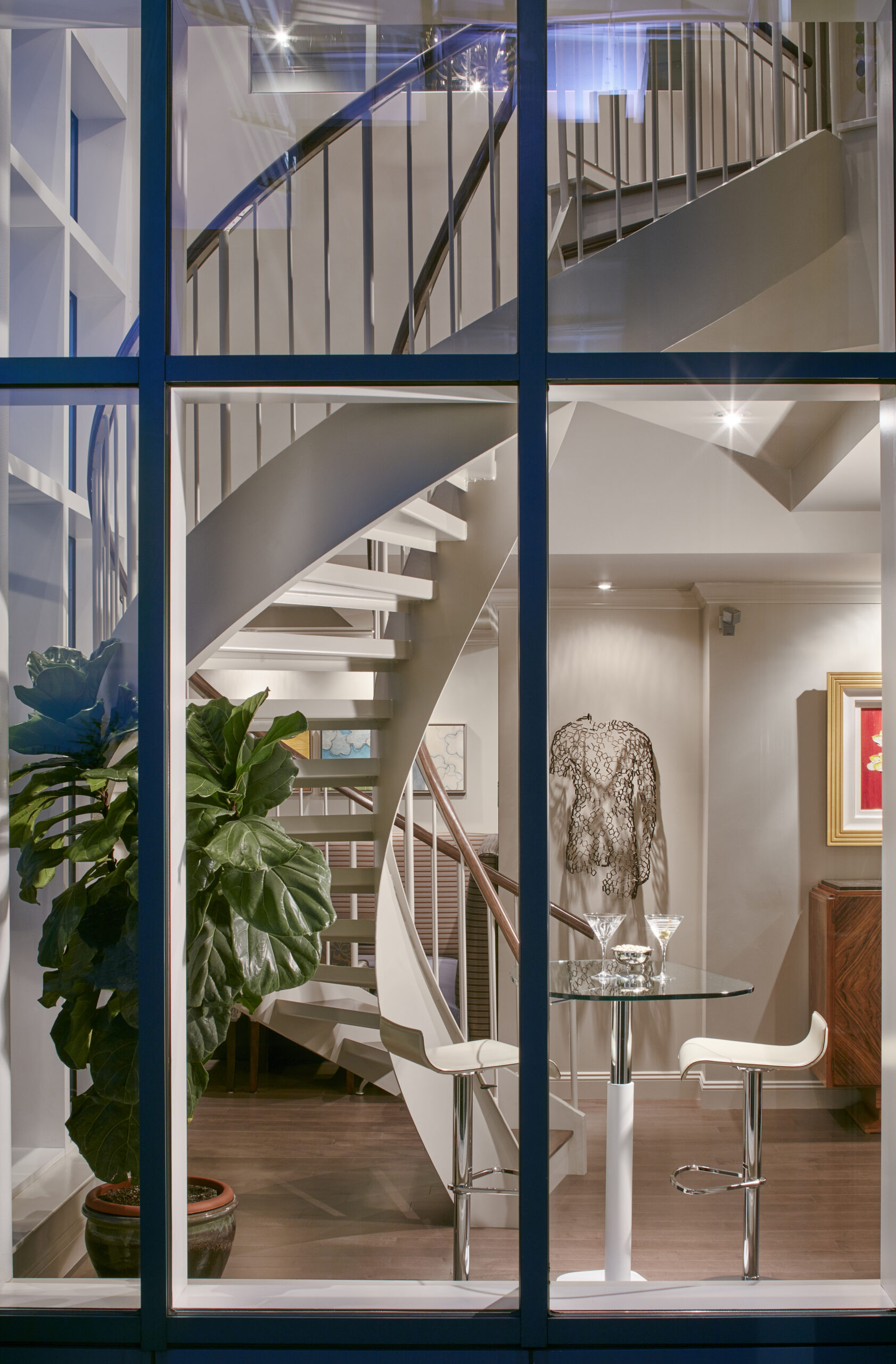 Modern luxury interior design with a spiral staircase, glass table and two white stools, a potted plant, and wall art, all elegantly arranged by an interior designer in Massachusetts, viewed through a large window.