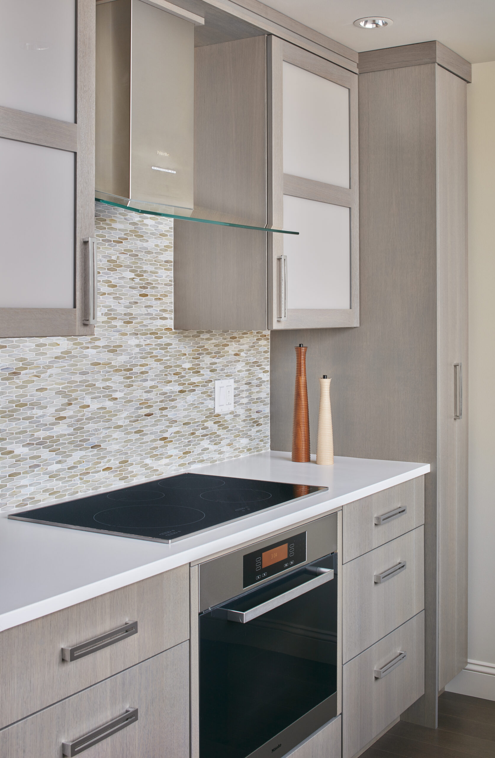 Modern kitchen with light wood cabinets, electric cooktop, built-in oven, mosaic tile backsplash, and two tall decorative vases on the counter—a perfect example of luxury interior design by an interior designer in Massachusetts.