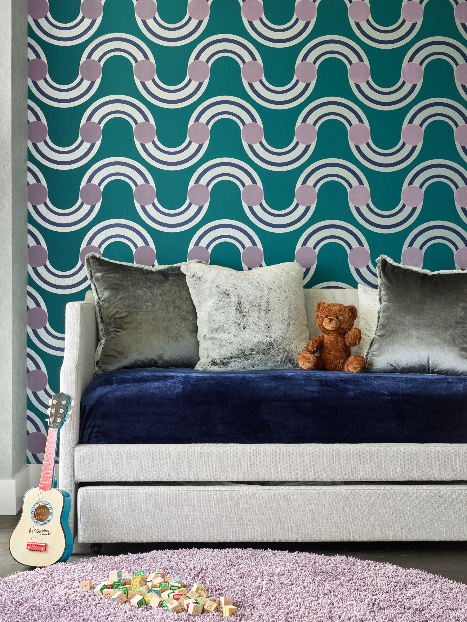 A child's room with a teal geometric wave wallpaper, a grey daybed with velvet cushions and a teddy bear, a toy guitar, alphabet blocks, and a lilac rug—showcasing the touch of an expert Interior Designer Massachusetts.