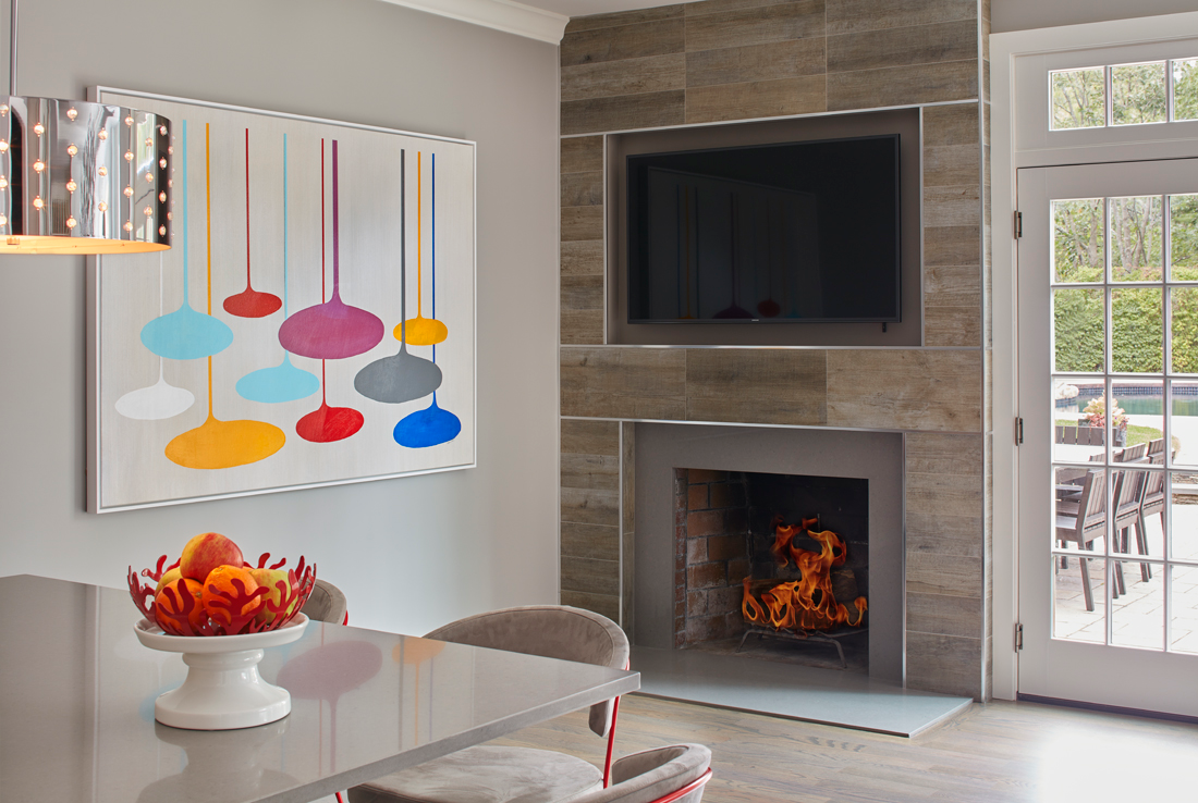 Modern living room with a wall-mounted TV above a lit fireplace, colorful abstract art, a dining table, and a fruit bowl centerpiece—an elegant example of Luxury Interior Design by an Interior Designer in Massachusetts.