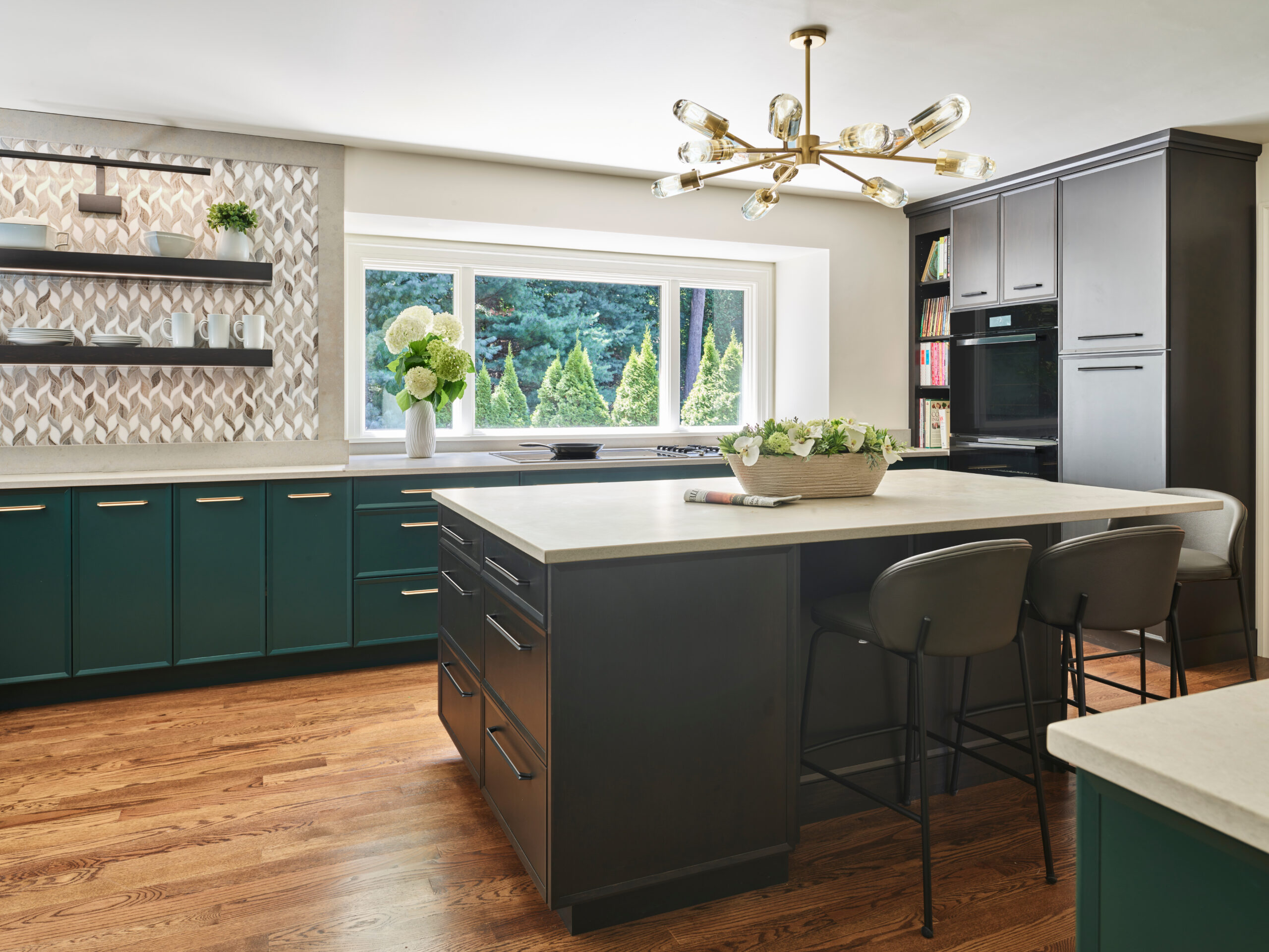 Modern kitchen with green cabinets, central island seating, large window, built-in oven, open shelves, decorative backsplash, and wood flooring—an example of Luxury Interior Design by a leading Interior Designer in Massachusetts.