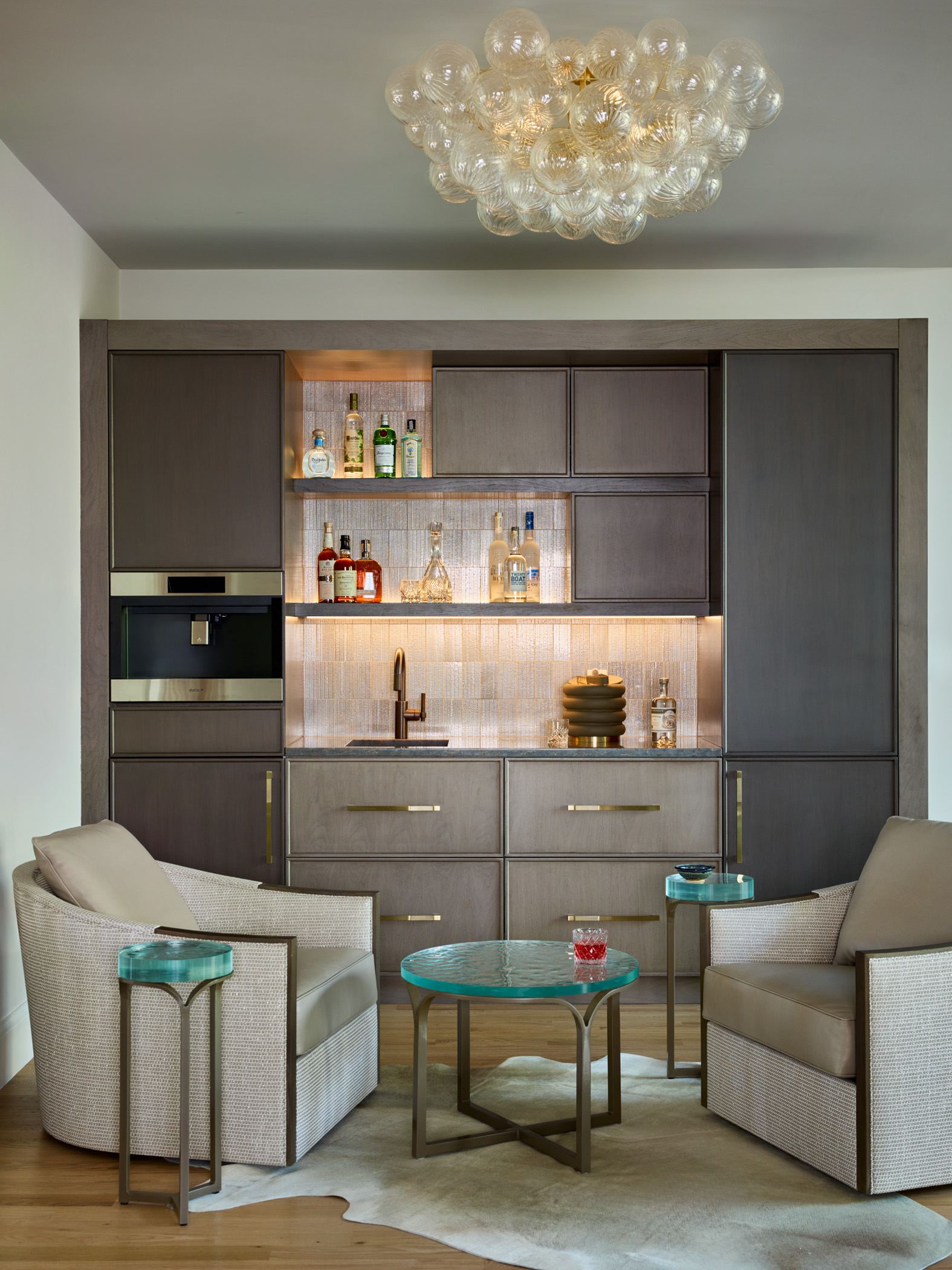 Modern kitchenette with gray cabinets, built-in bar, and open shelves displaying glassware and liquor bottles. Two beige armchairs and small green-topped tables add a touch of Luxury Interior Design to the inviting space.