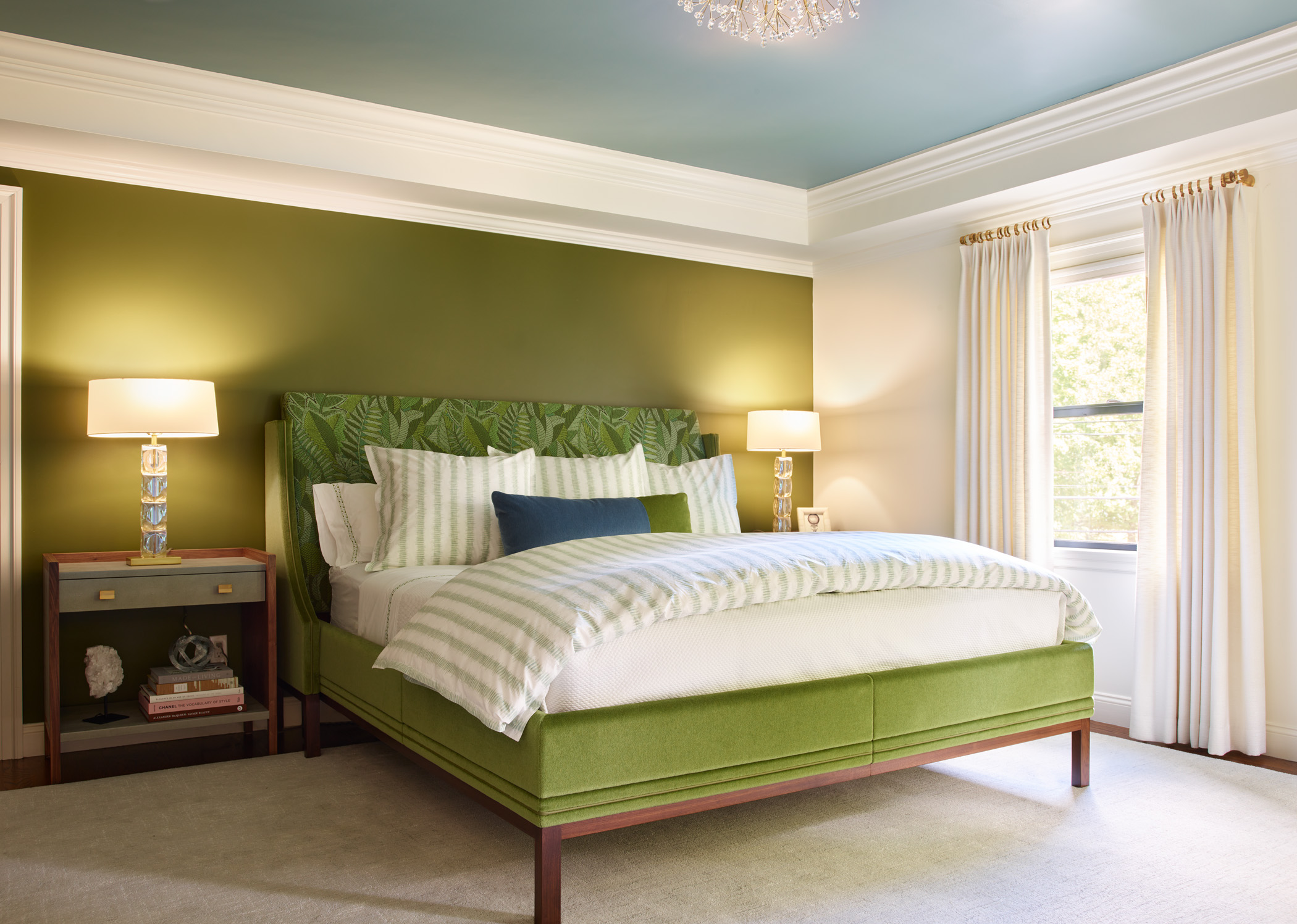 A bedroom with a green upholstered bed, striped bedding, green accent wall, bedside tables with lamps, large window with white curtains, and a light blue ceiling showcases luxury interior design by an Interior Designer in Massachusetts.
