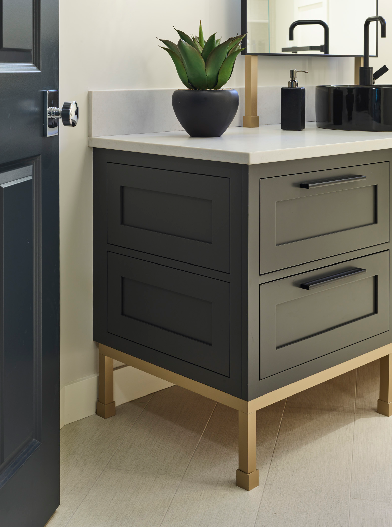 Modern bathroom vanity with black drawers, gold legs, and a white countertop—a perfect touch of luxury interior design—features a potted plant, soap dispenser, and square mirror against a light wall and tile floor.
