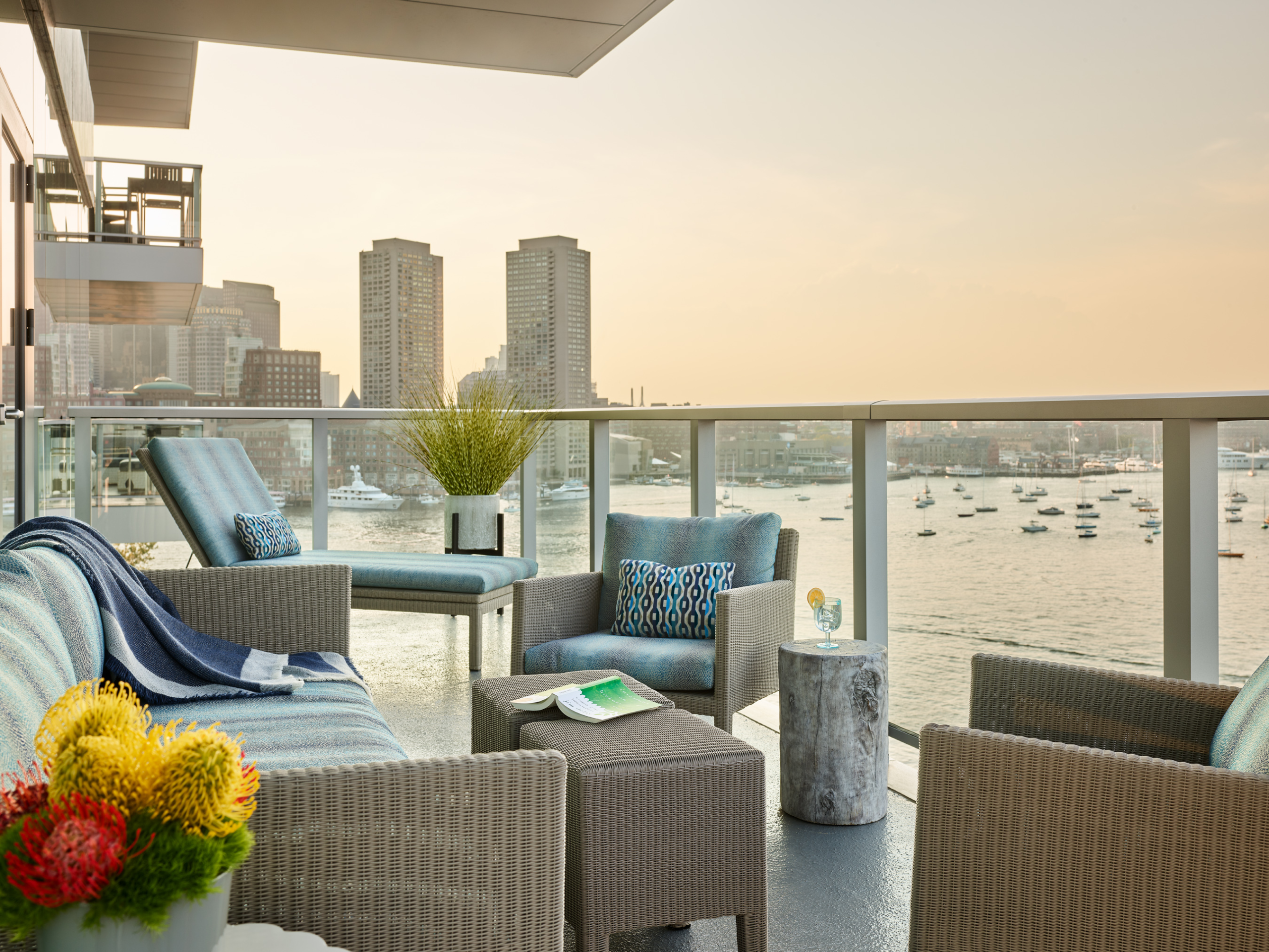 Experience a modern outdoor seating area curated with Luxury Interior Design on a balcony overlooking the harbor, boats, and city skyline at sunset—an elegant retreat inspired by top Interior Designer Massachusetts.