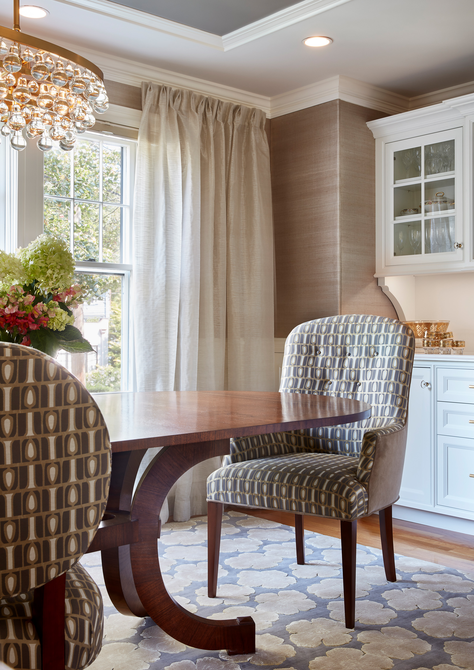 A dining room with a wooden table, patterned upholstered chairs, a floral arrangement, sheer curtains, a window, a chandelier, and white cabinetry with glass doors showcases luxury interior design.