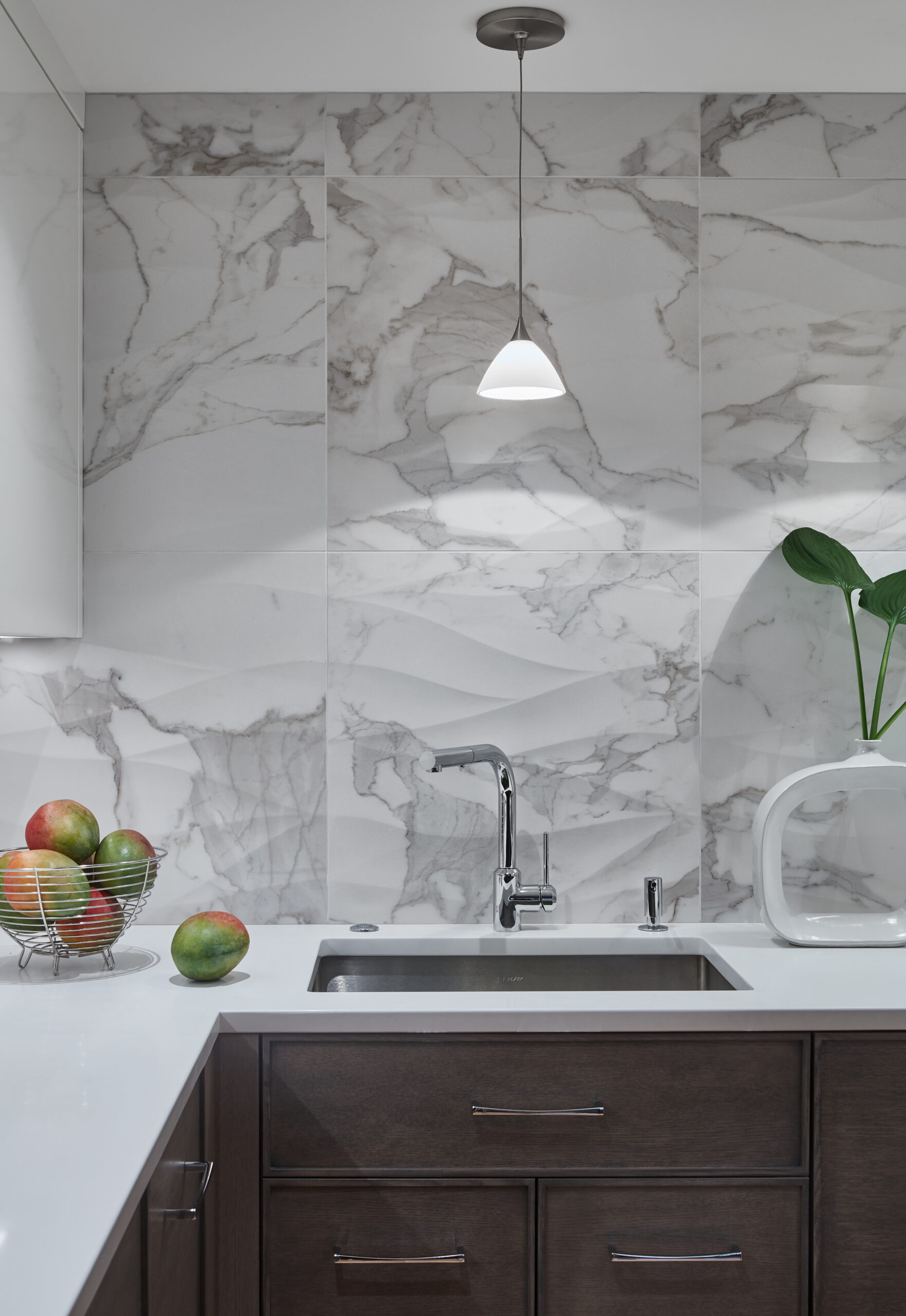 Modern kitchen with marble backsplash, dark wood cabinets, a stainless steel sink, a pendant light, a bowl of mangoes, and a white vase with green leaves reflects the essence of luxury interior design.