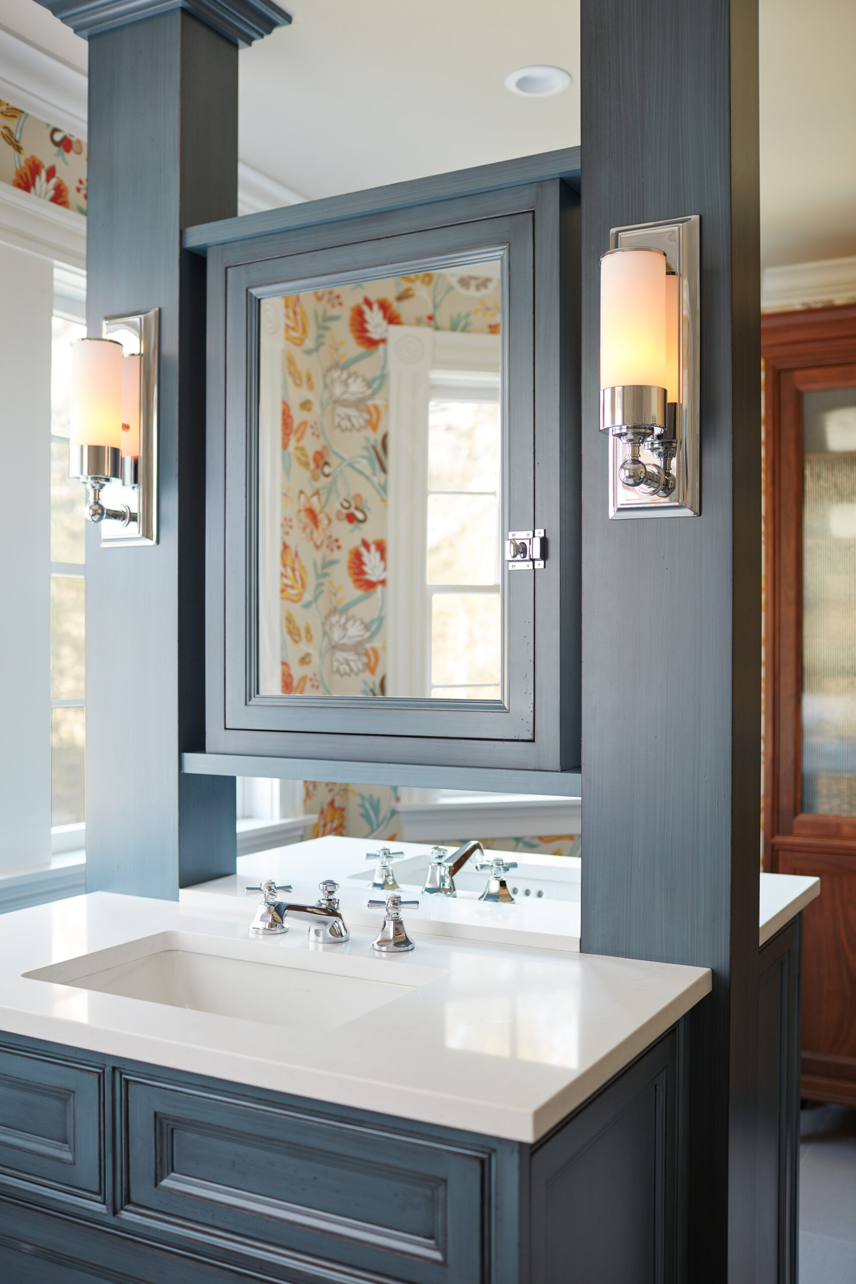 A blue bathroom vanity with a white countertop, mirrored cabinet, chrome faucet, and wall-mounted light fixtures exudes Luxury Interior Design against floral wallpaper in the background.