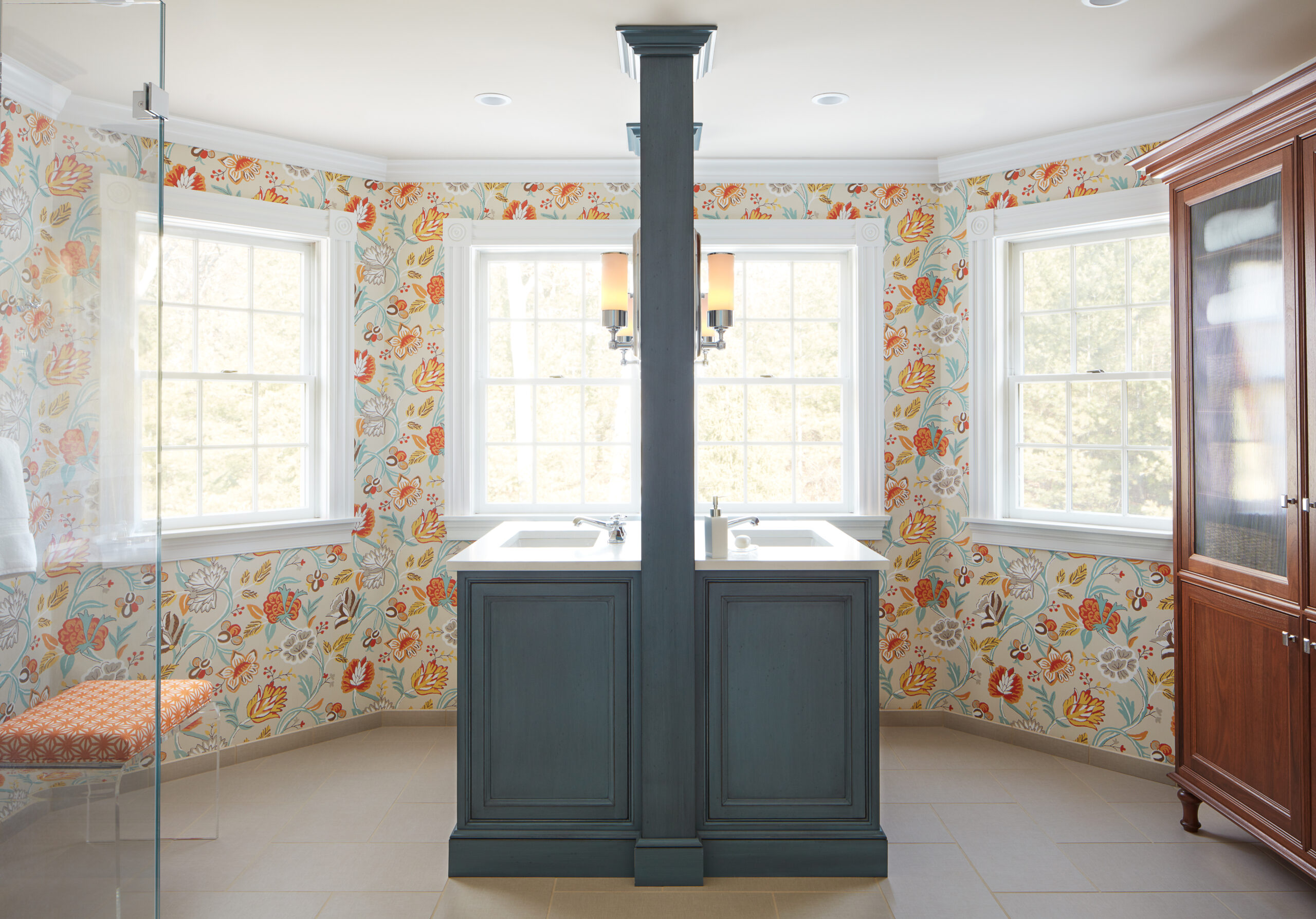 A bathroom designed with floral wallpaper, luxury interior design touches, two windows, a double sink vanity with blue cabinets, and a wooden wardrobe—perfect for those seeking sophistication.