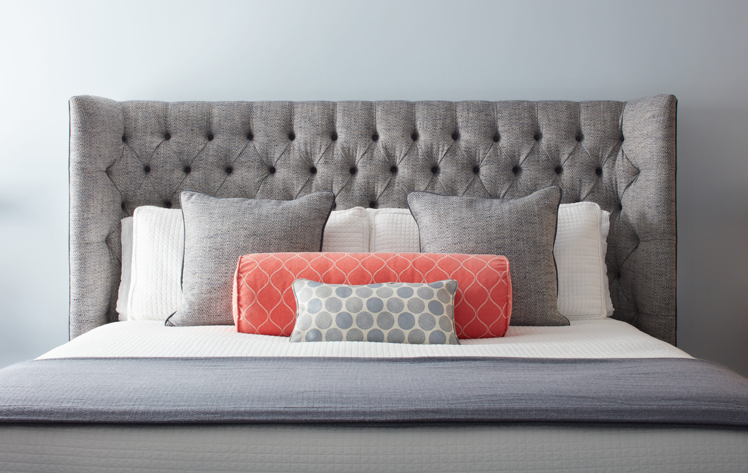 A neatly made bed with a tufted gray headboard, gray pillows, white bedding, and two accent pillows in coral and gray patterns reflects the elegance of luxury interior design.