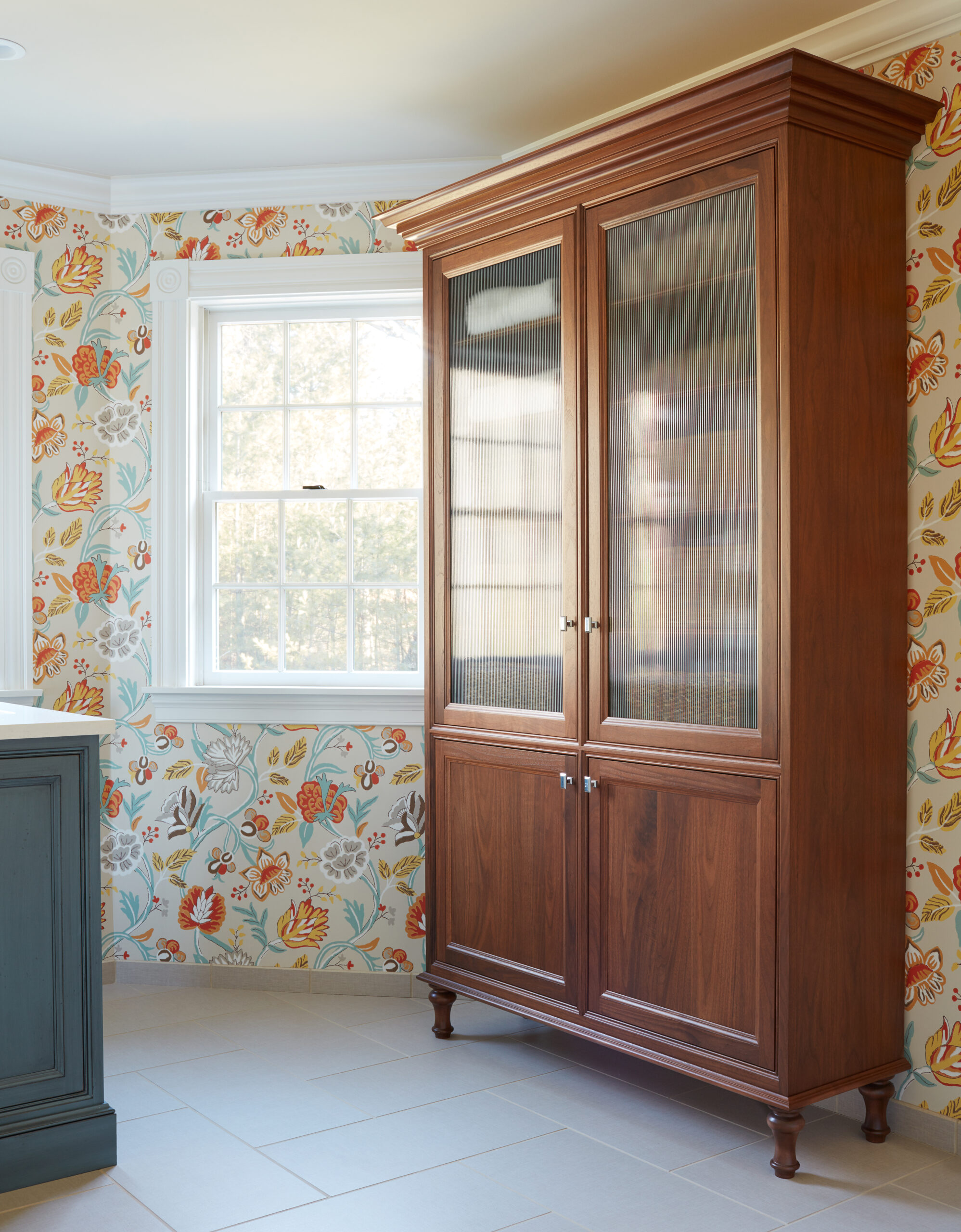 A tall wooden cabinet with glass upper doors stands against a wall with floral wallpaper near a window in a brightly lit room, reflecting the elegance of luxury interior design.