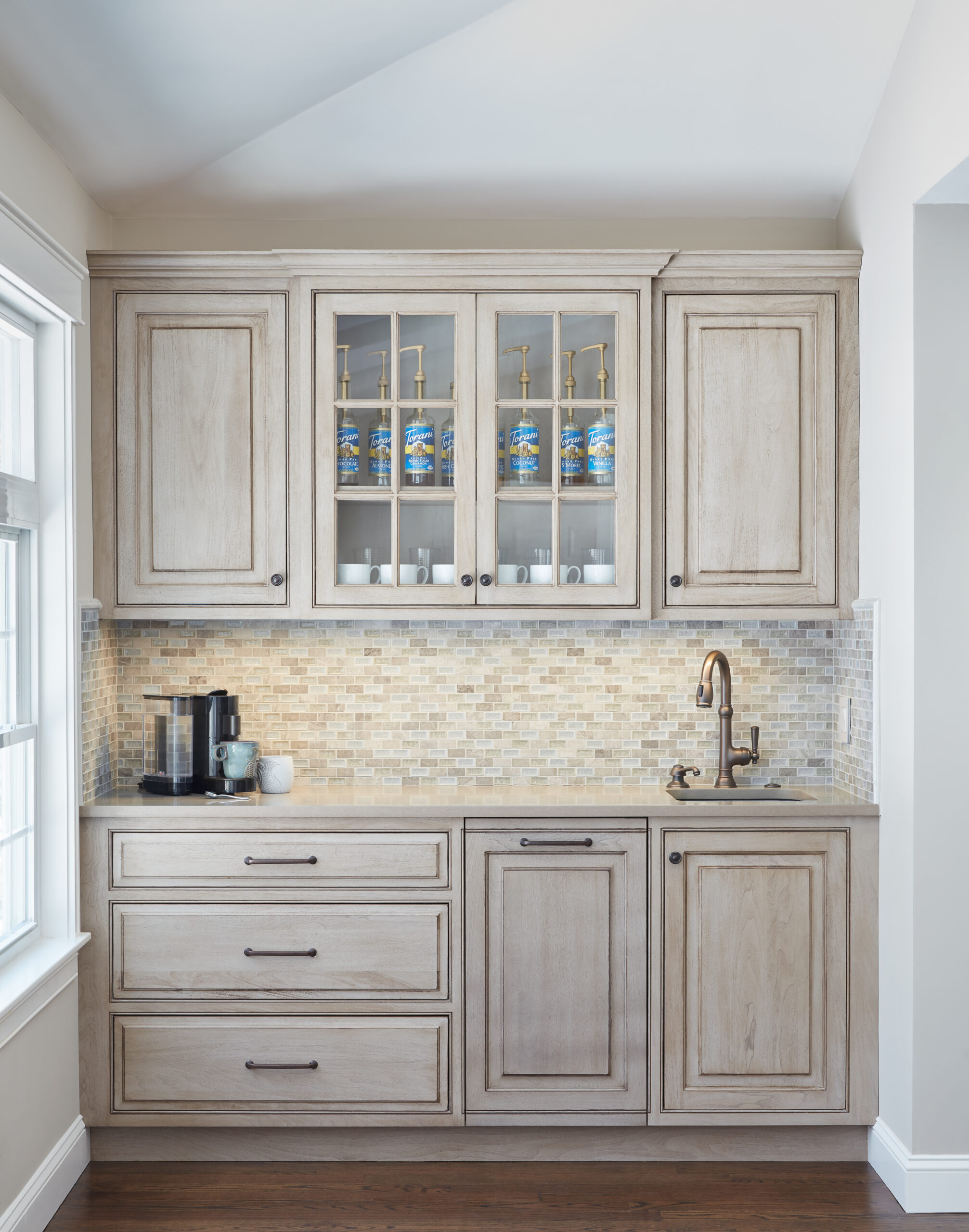 Light wood kitchen cabinets with a glass-front upper section displaying cups and syrup bottles, a coffee machine, a small sink with a bronze faucet, and a tiled backsplash—perfect for those who appreciate luxury interior design.