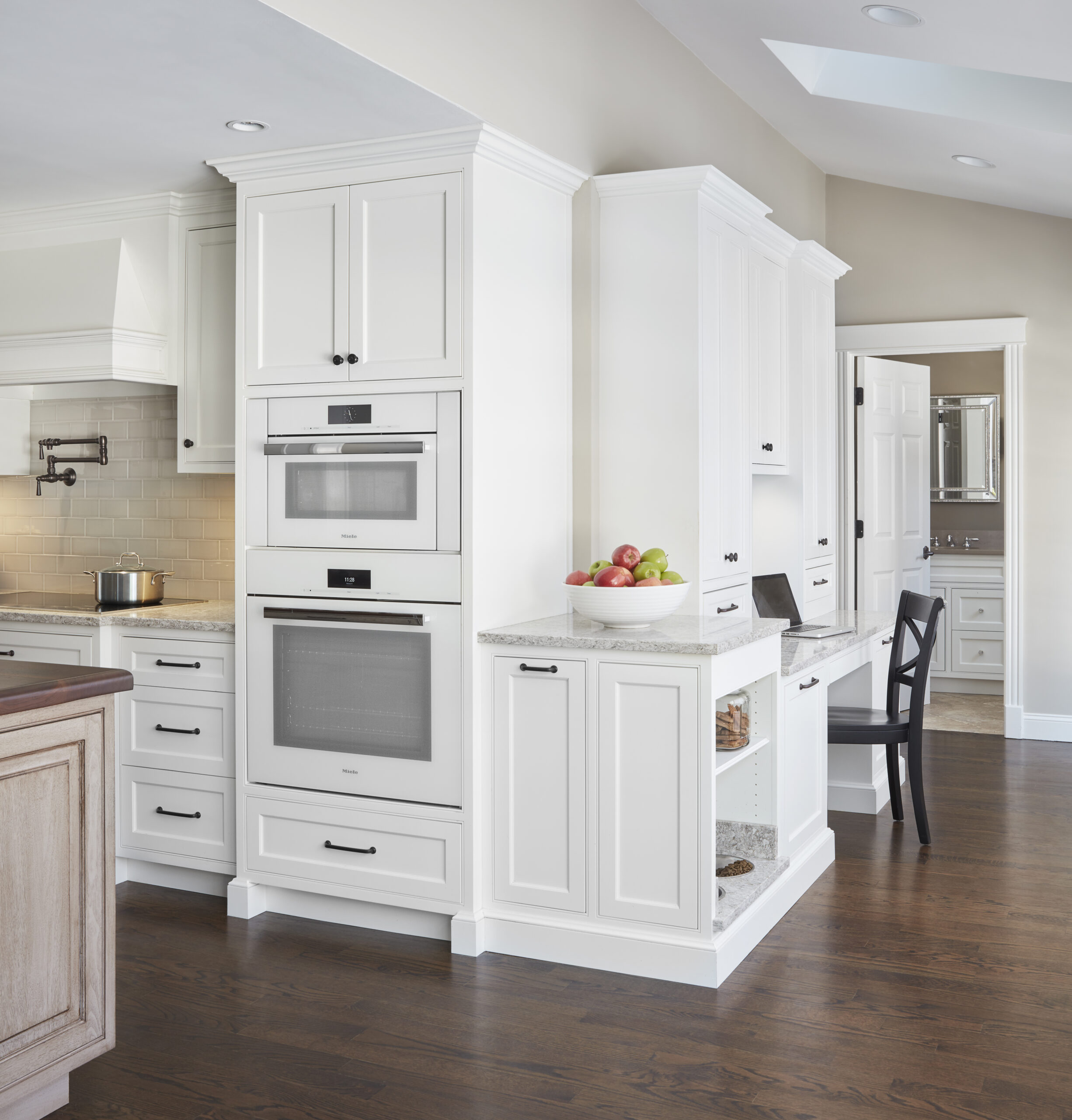 A modern kitchen with white cabinetry, built-in ovens, a desk area with a chair, a bowl of apples on the counter, and dark hardwood floors showcases luxury interior design at its finest.