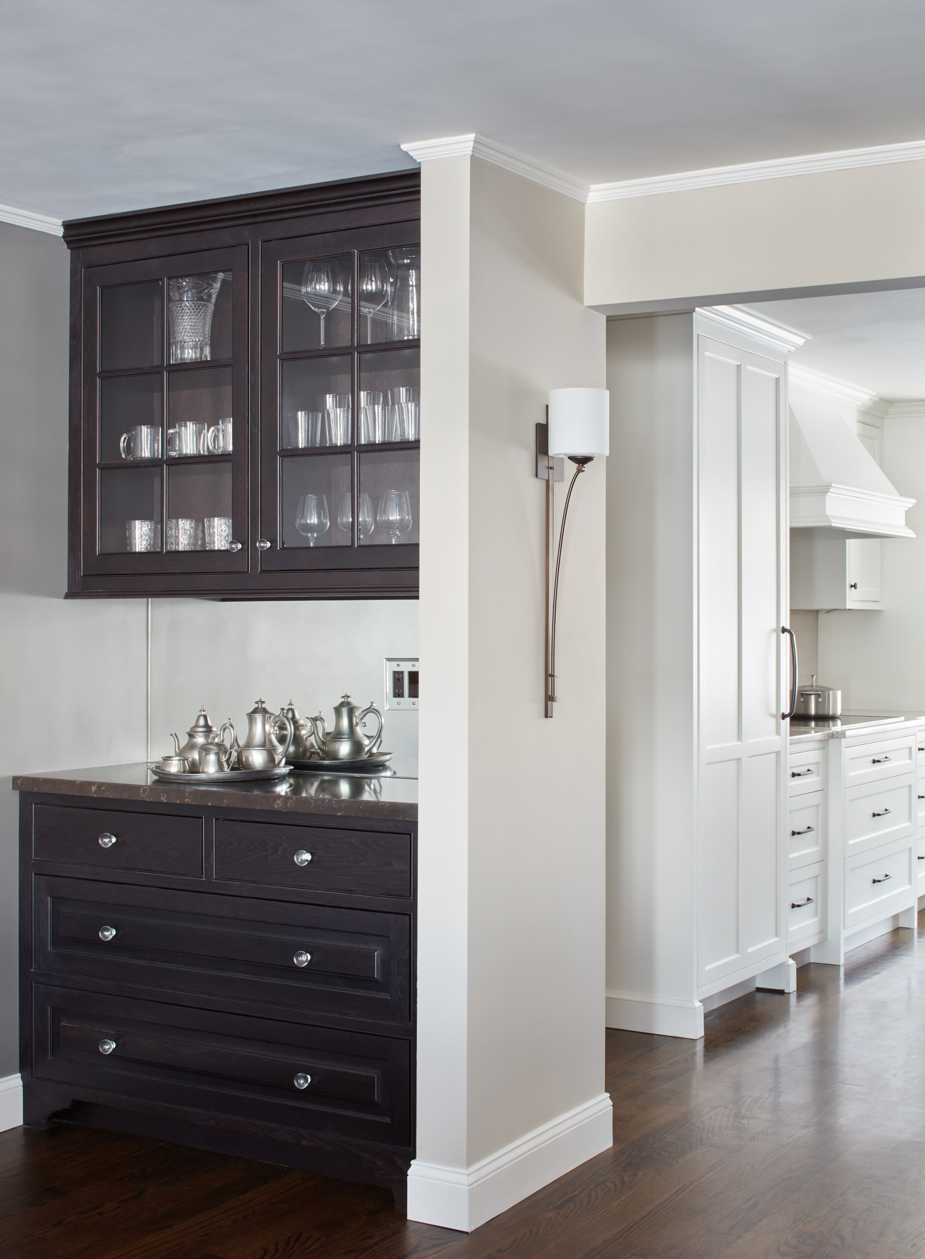 A dark wood cabinet with glass doors displays glassware above drawers holding silver serving pieces; a modern wall sconce is mounted beside it, giving this luxury interior design a refined touch, with a white kitchen visible in the background.