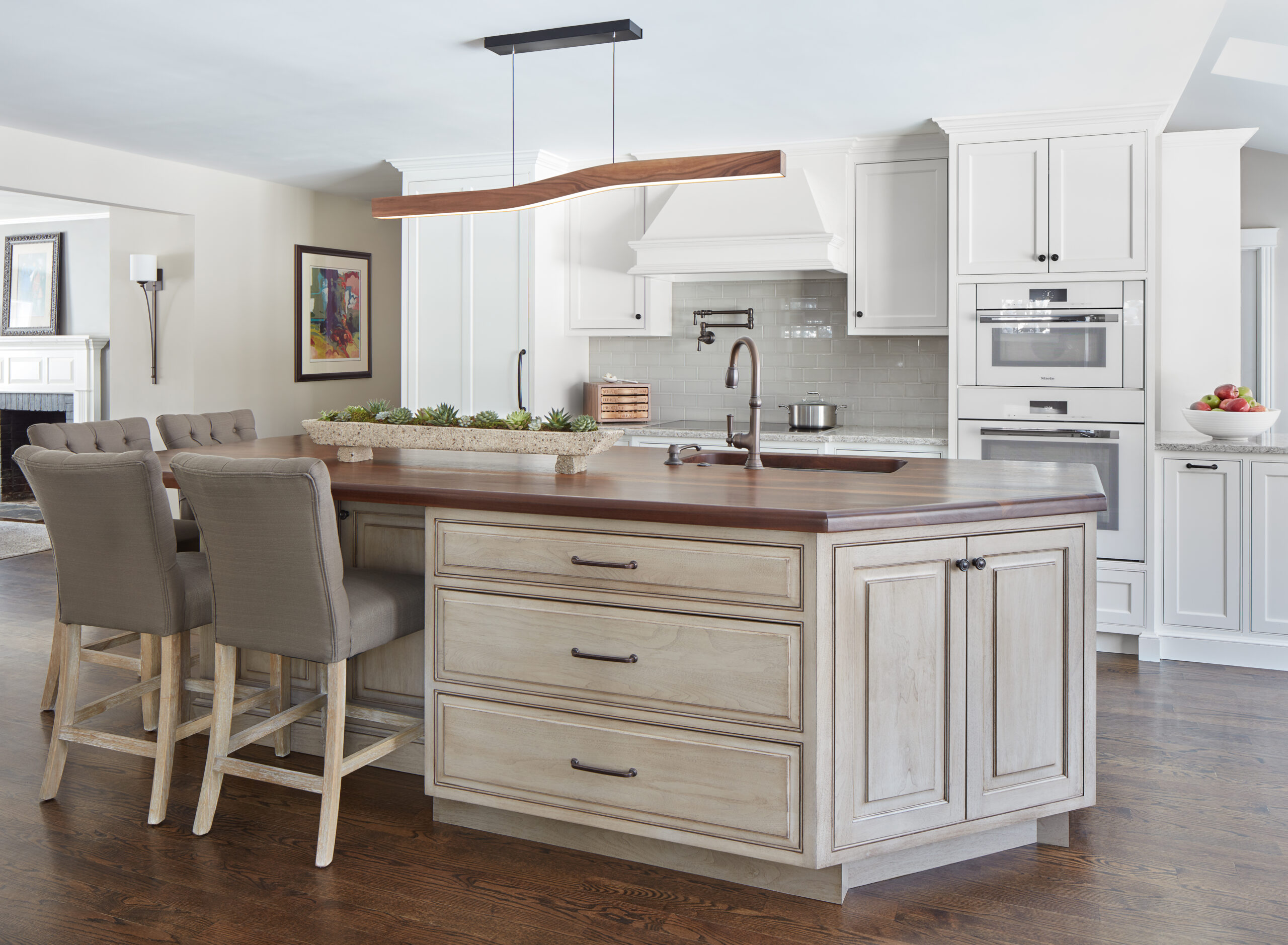 Modern kitchen with a wooden island, three upholstered chairs, white cabinetry, stainless steel appliances, and a wave-shaped pendant light—showcasing the elegance of Luxury Interior Design by an expert Interior Designer Massachusetts.