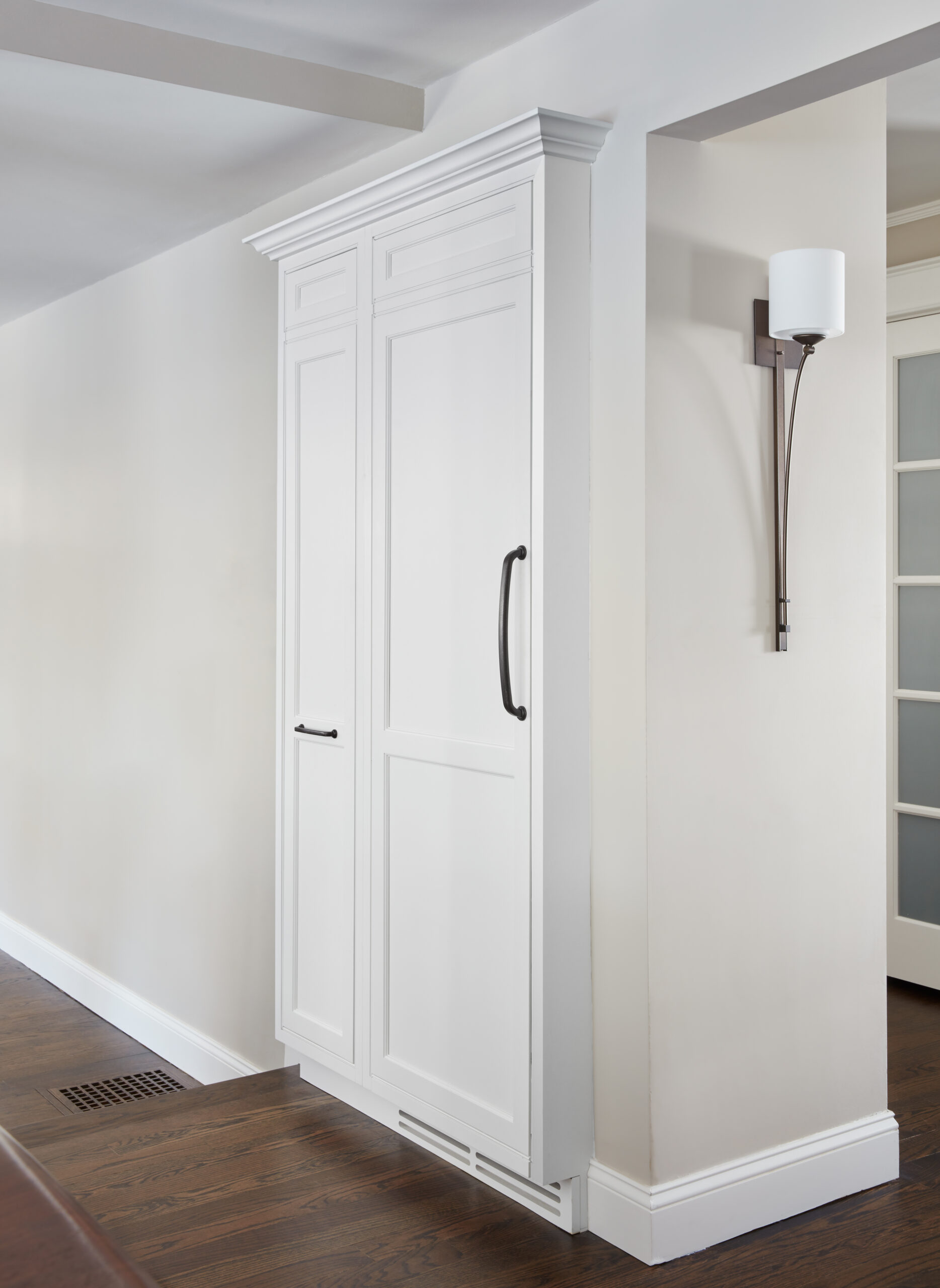Tall white built-in cabinet with two doors and black handles, styled by a luxury interior design expert, set against a beige wall next to a sconce and glass-paneled door on a dark wood floor.