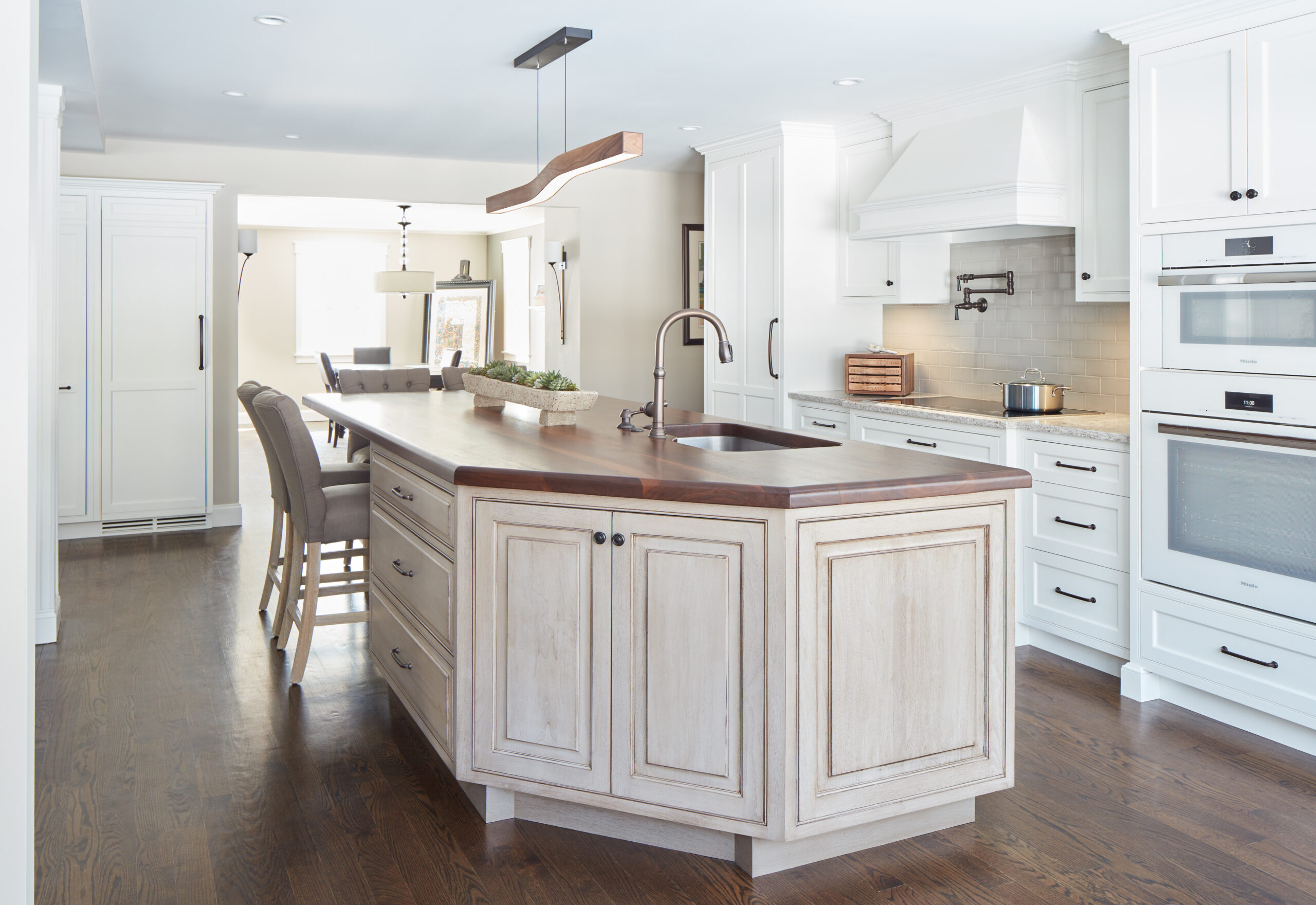Modern kitchen with white cabinets, a large wooden island with sink, built-in oven, and stovetop—an inviting space that showcases luxury interior design by a leading interior designer in Massachusetts. Hardwood floors and neutral decor complete the look.