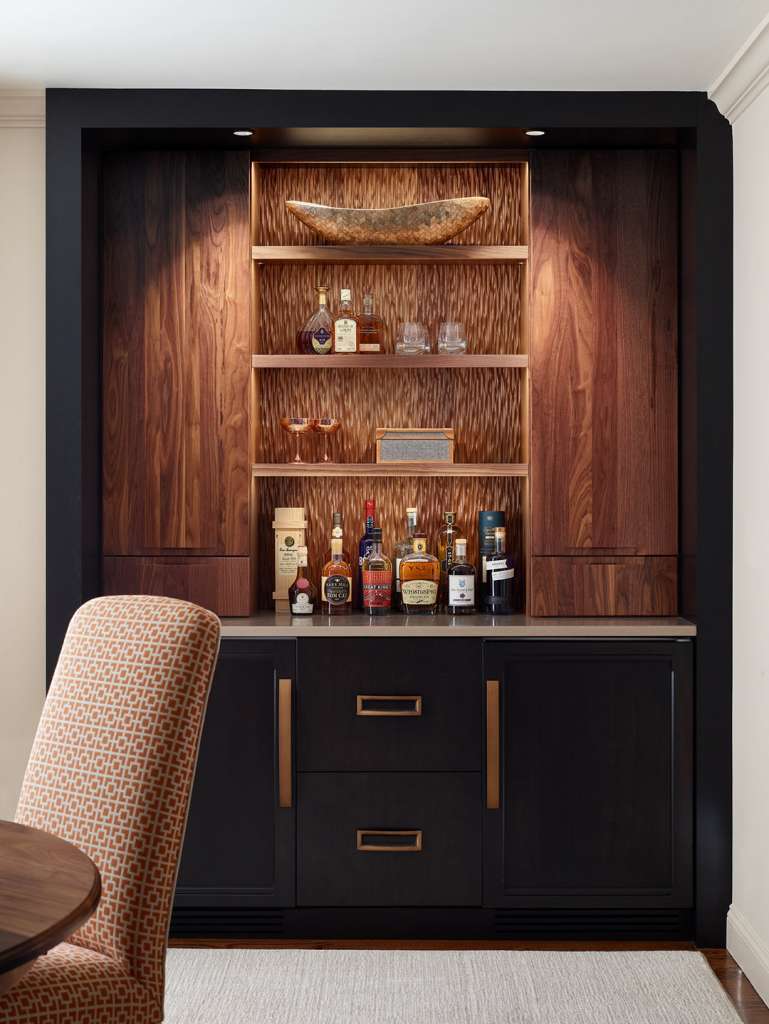 A built-in home bar with dark wood cabinets, open wooden shelves displaying bottles of liquor, glassware, and a decorative bowl, set against a textured wood backdrop—a stunning example of luxury interior design.
