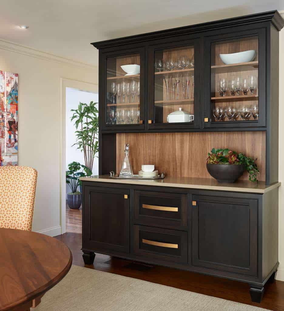 A black wooden hutch with glass doors displays glassware and dishes, while a countertop holds a tray, bowl, and decorative items—perfect for a luxury interior design touch inspired by an Interior Designer Massachusetts.
