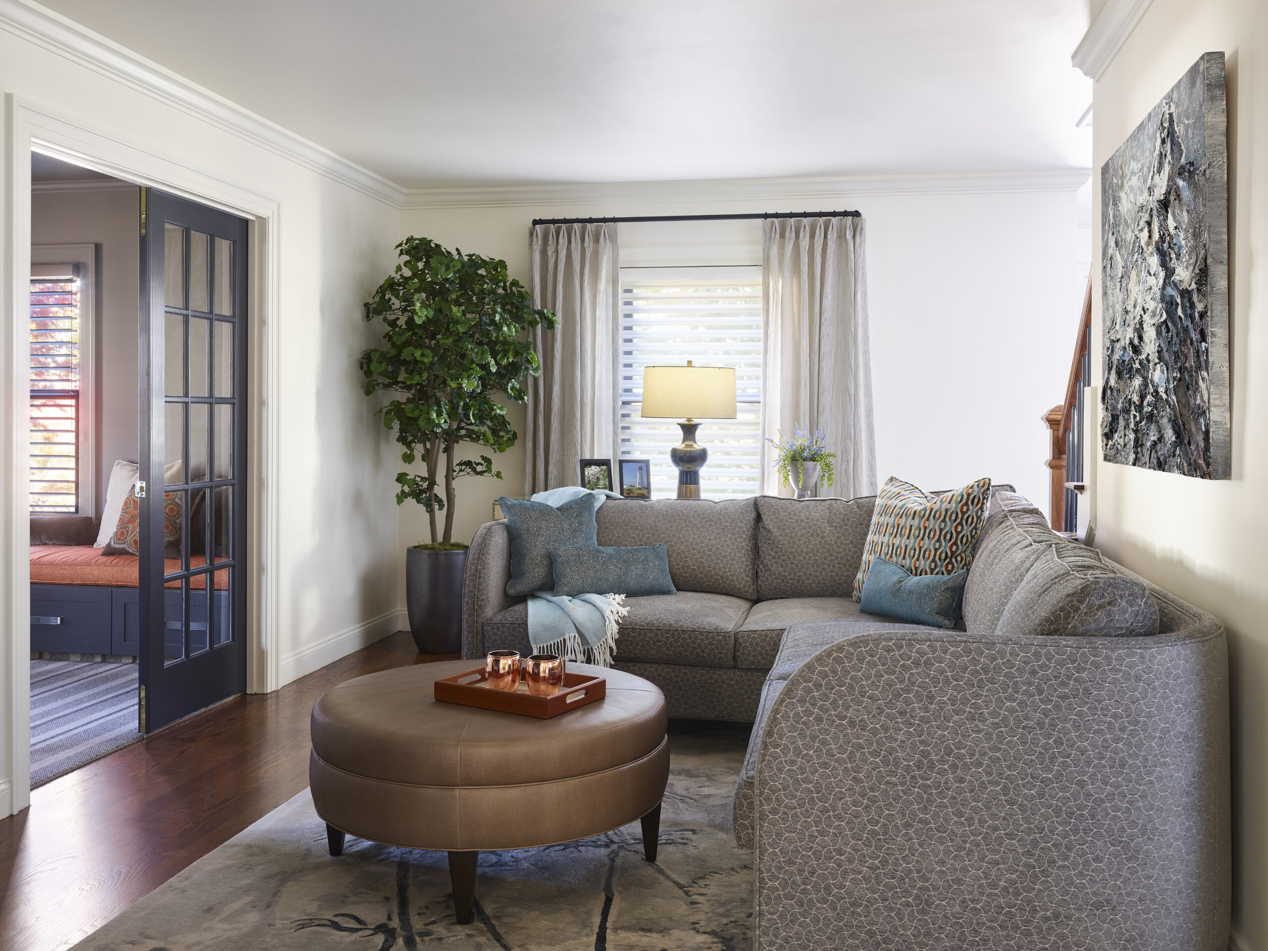 A living room with a gray sectional sofa, round brown ottoman, floor plant, table lamp, and abstract wall art exudes luxury interior design, featuring double doors that lead to another room.