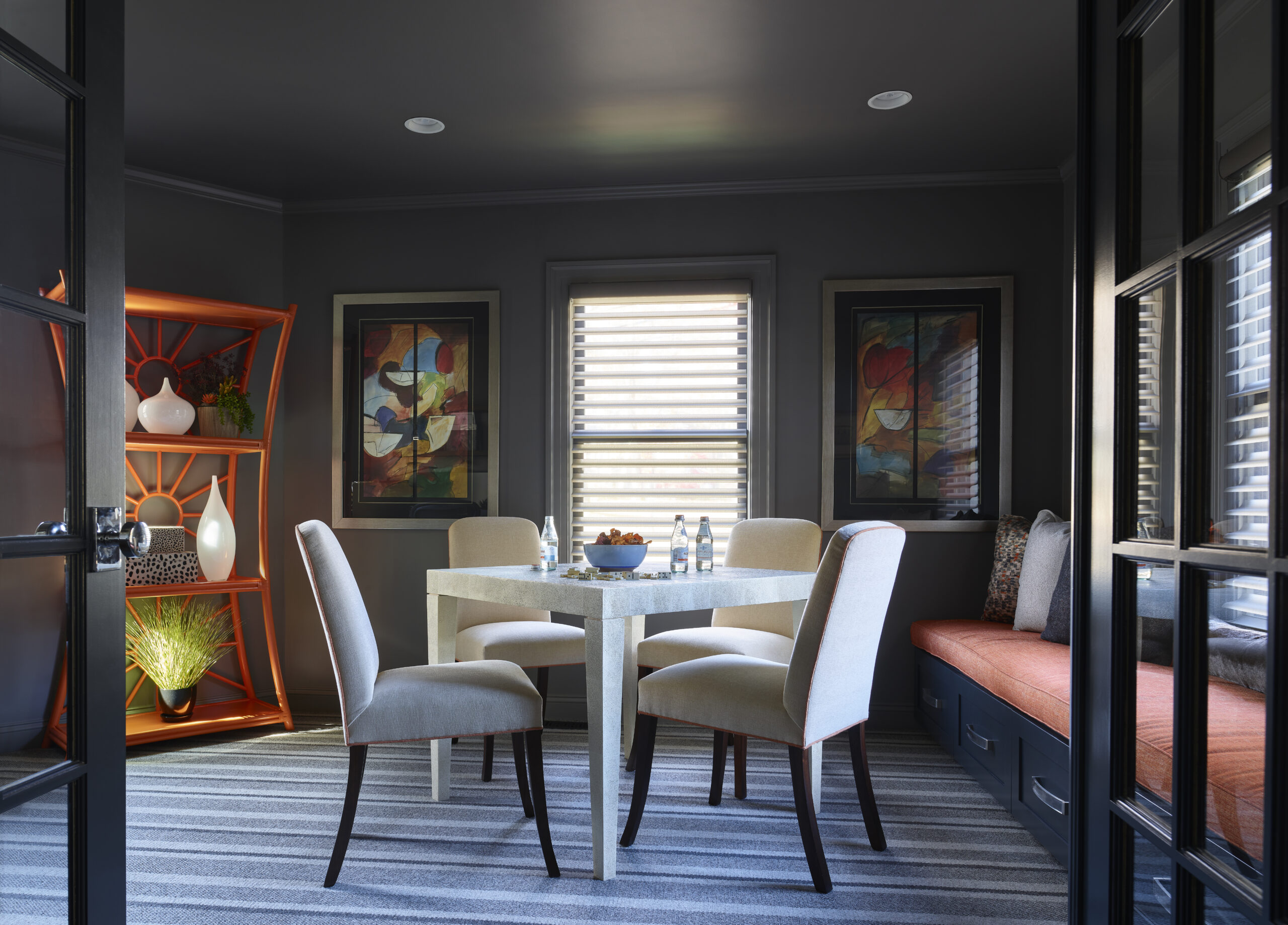 Modern dining nook designed by an Interior Designer Massachusetts, featuring a square table, four upholstered chairs, built-in bench with orange cushion, striped rug, abstract art, and an orange shelving unit with decor items for a touch of luxury interior design.