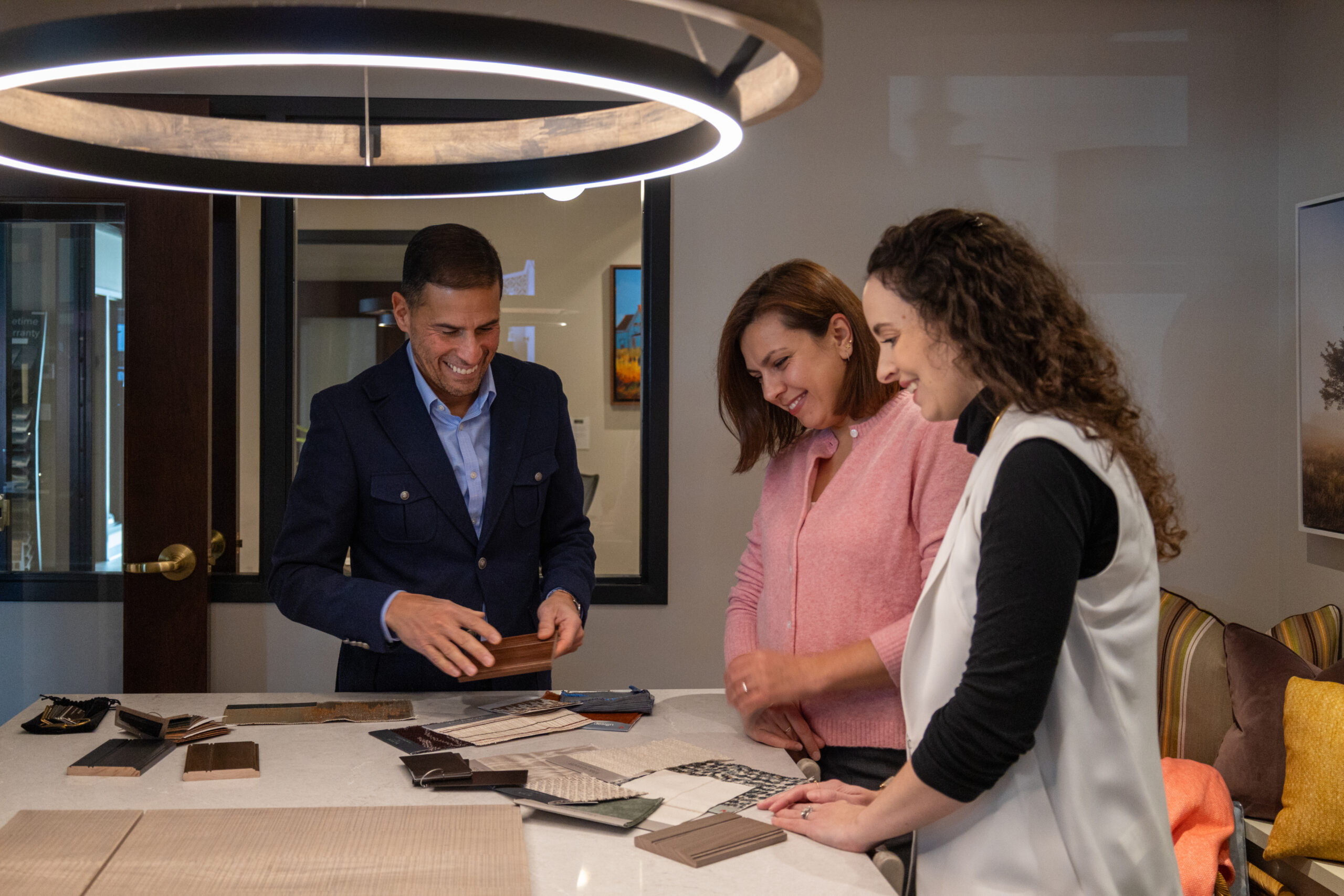 Three people stand around a table, examining and discussing various fabric and material samples under a circular ceiling light—an inspiring moment in luxury interior design guided by an expert interior designer from Massachusetts.