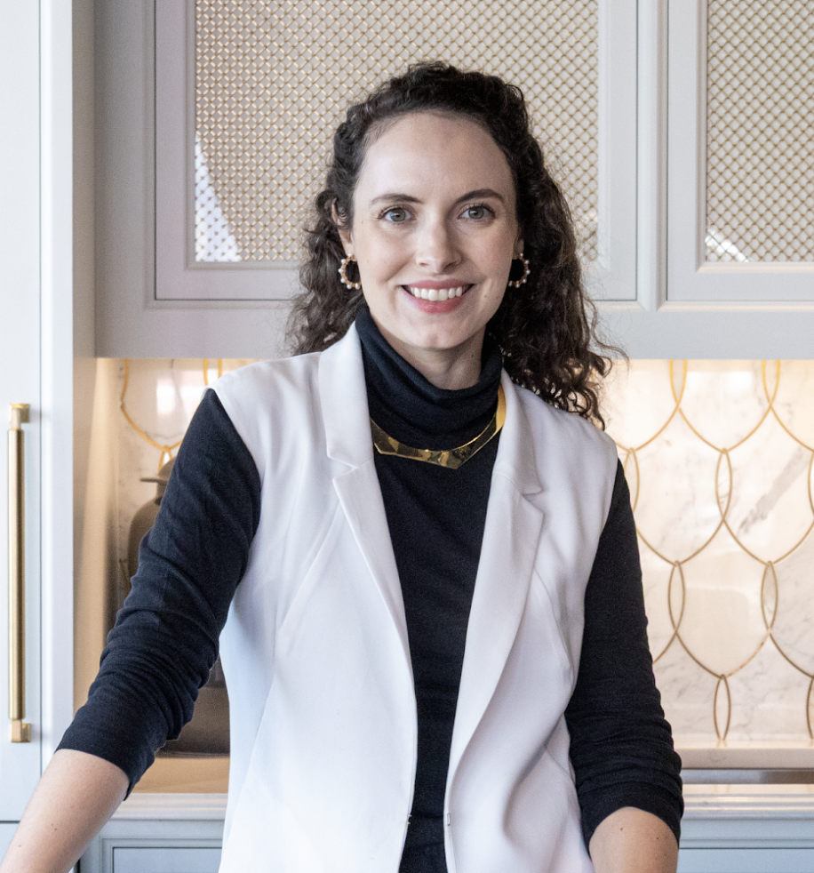 Woman with curly hair, an interior designer in Massachusetts, wears a white vest over a black turtleneck and stands in a modern kitchen with light cabinets and gold accents, smiling at the camera, showcasing luxury interior design.