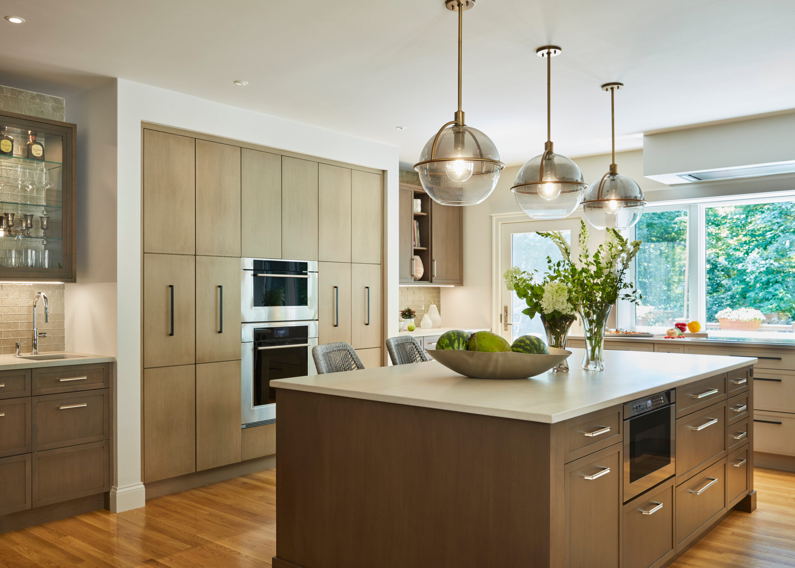 Modern kitchen with wooden cabinets, a large island with built-in stove, pendant lights, bar stools, and a window overlooking greenery—perfectly showcasing luxury interior design at its finest.