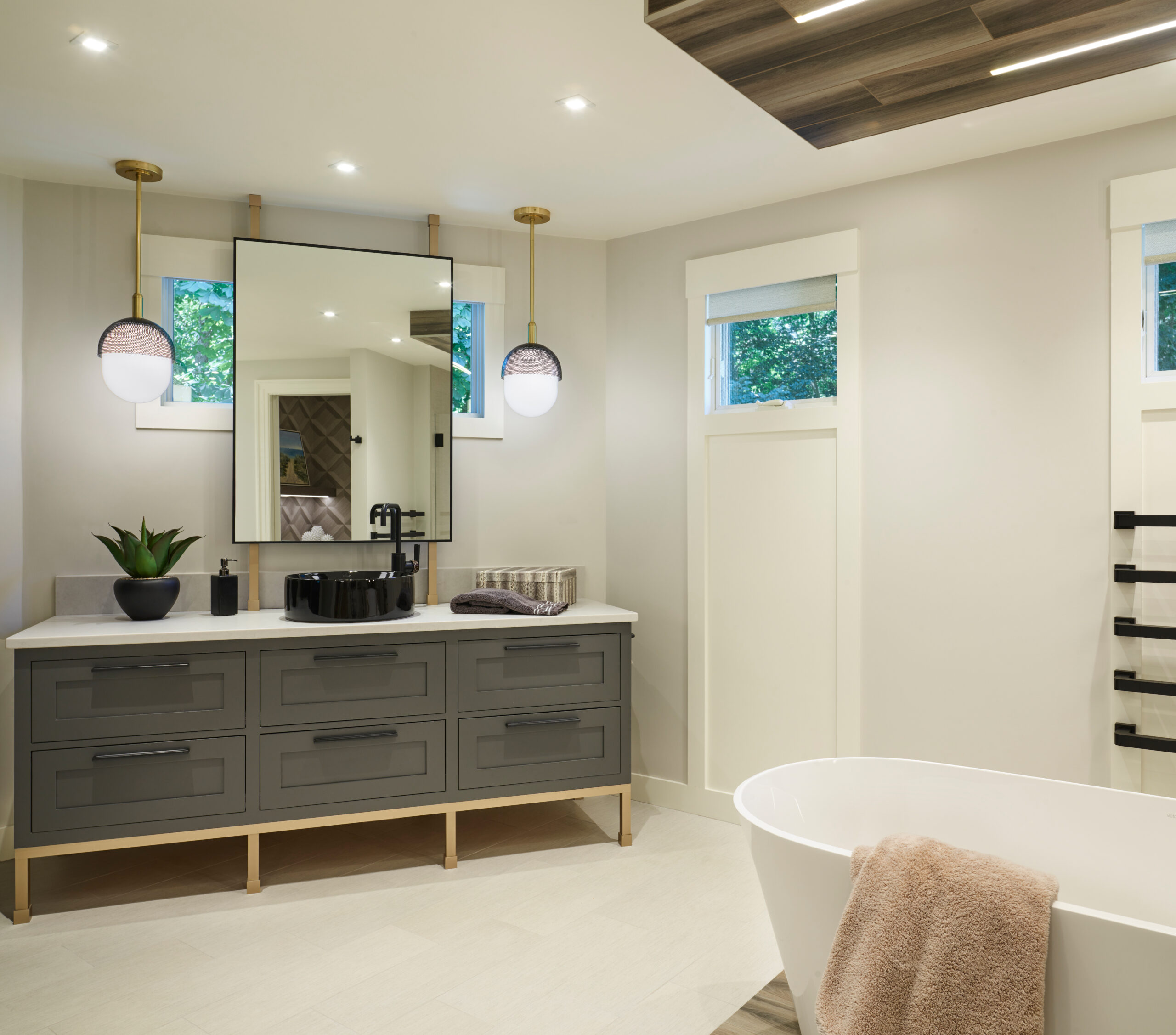 Modern bathroom with a gray vanity, two pendant lights, a rectangular mirror, freestanding tub with a brown towel, and two small windows letting in natural light—perfect for showcasing luxury interior design by an expert interior designer in Massachusetts.