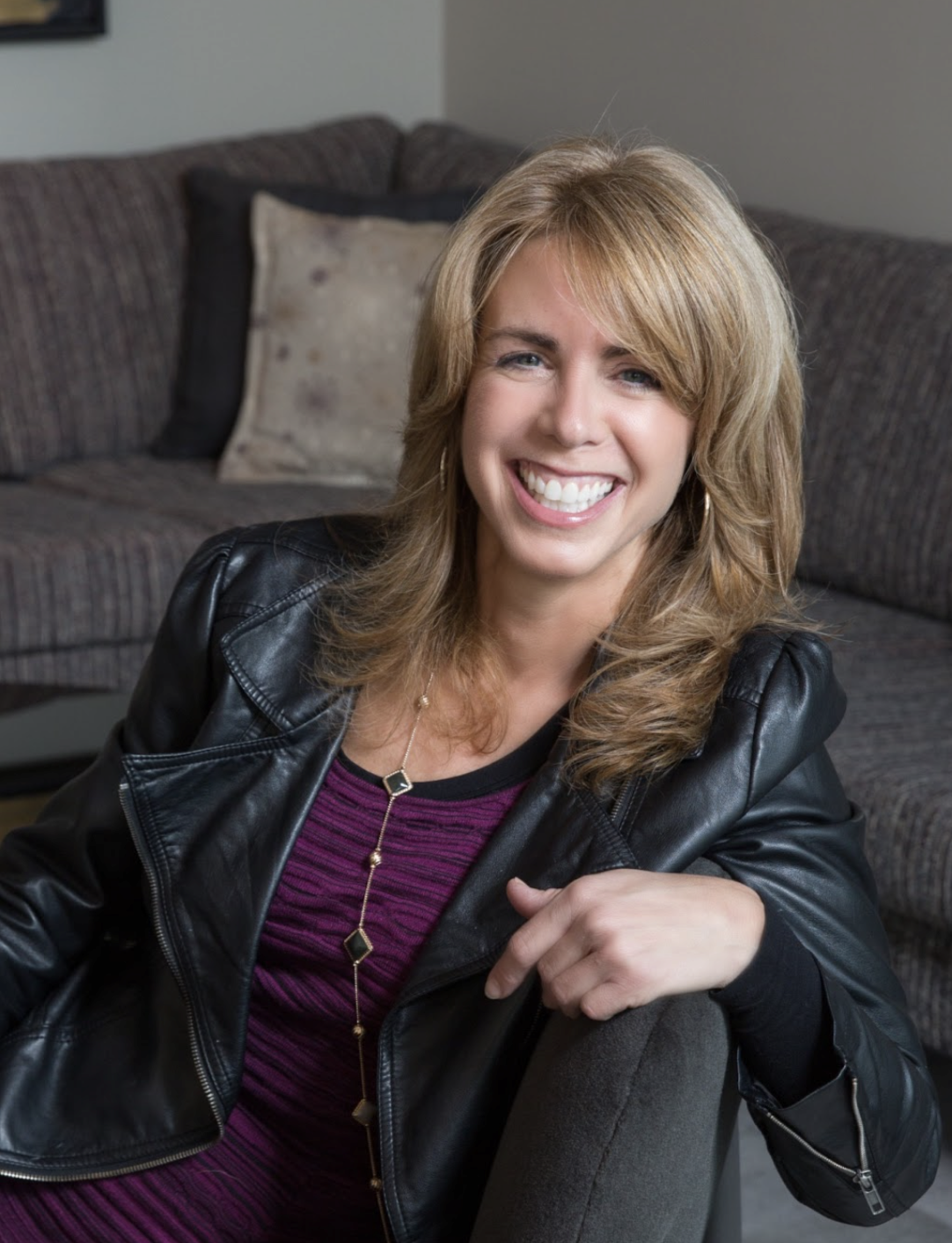 A woman with blonde hair, an Interior Designer from Massachusetts, sits on a sofa, smiling warmly as she wears a black leather jacket over a purple dress—an embodiment of luxury interior design style.